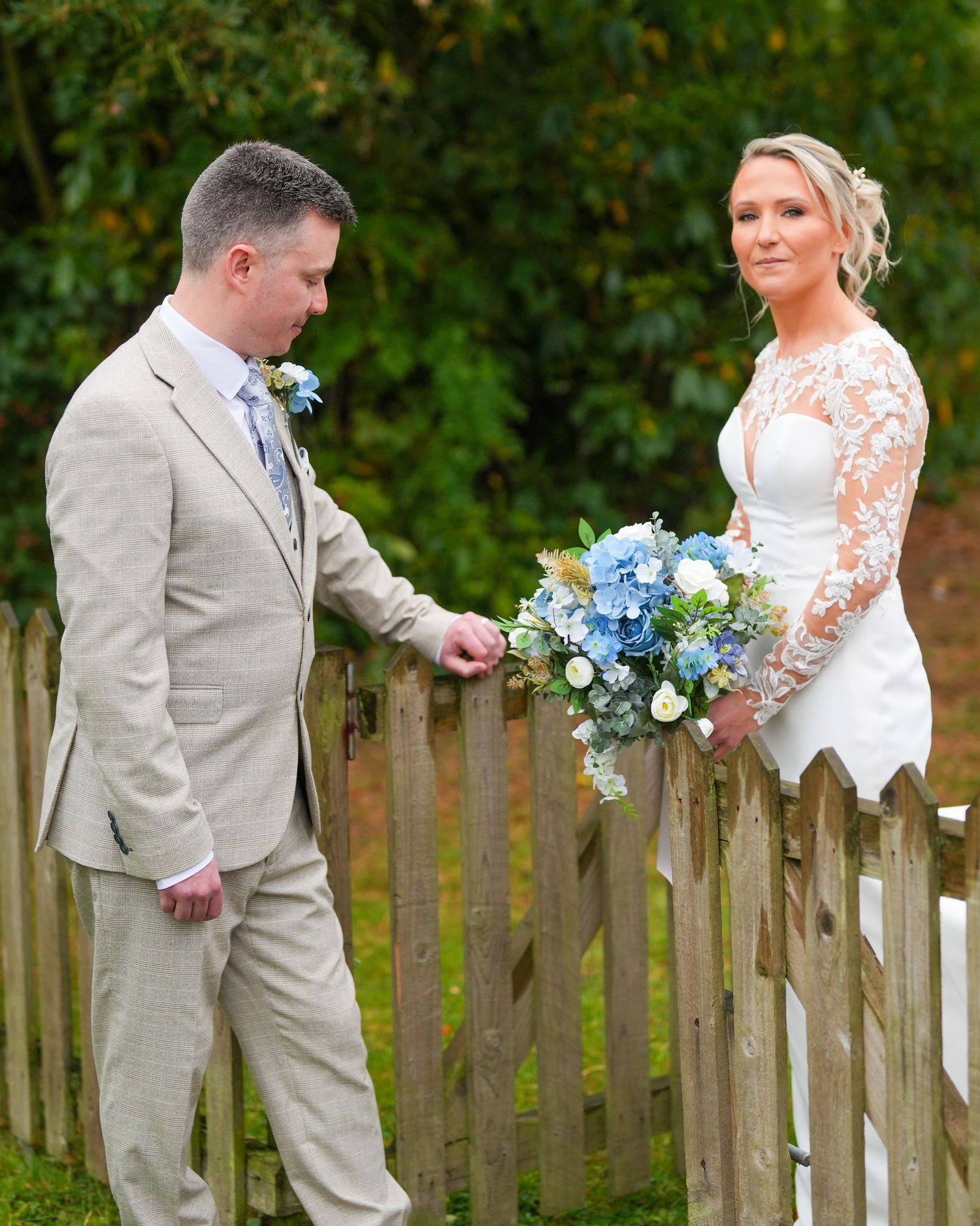 Bride in lace sleeve gown holds blue bouquet by wooden fence as groom opens gate. Dave Martin Photographer Northern Ireland