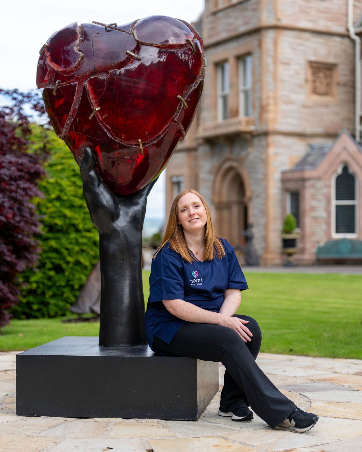 Young woman in navy Heart scrubs sitting beside a large red heart sculpture outside a historic stone building. Dave Martin Photography North