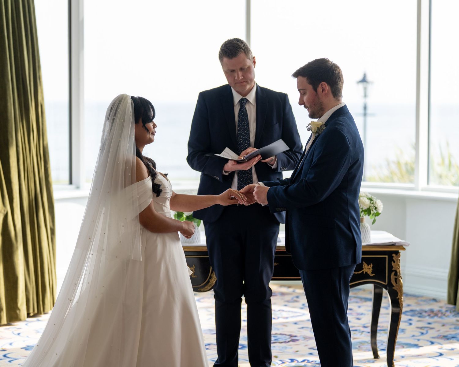Bride and groom exchange rings during indoor wedding ceremony with officiant reading from book by bright window. Dave Martin Photographer No