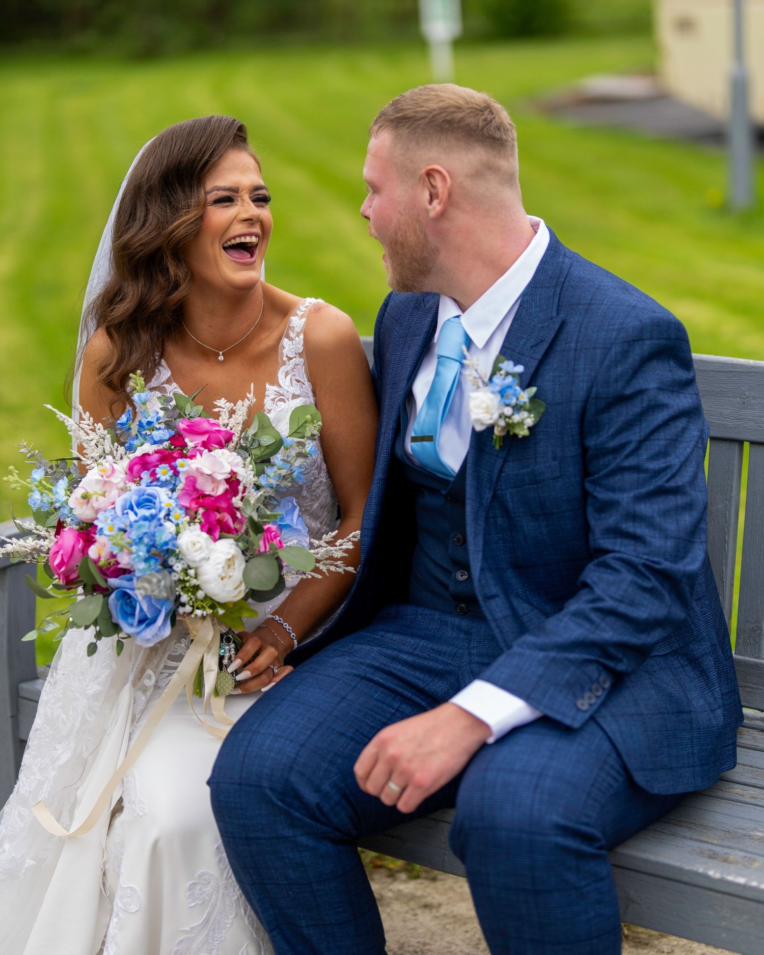 A joyful bride and groom in blue suit sit on bench laughing, bride holding colorful pink and blue wedding bouquet. By Dave Martin Photograph