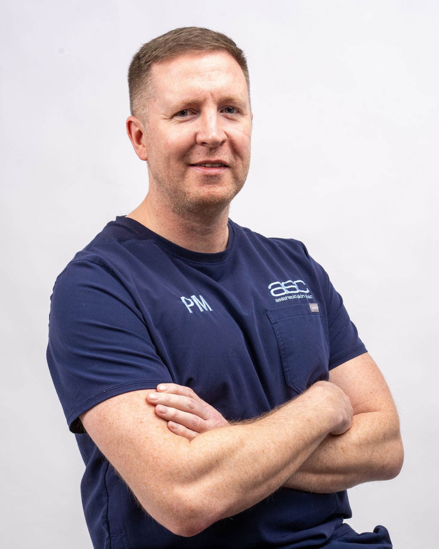 Professional male in navy scrubs with arms crossed, smiling confidently against white background. Dave Martin studio Belfast