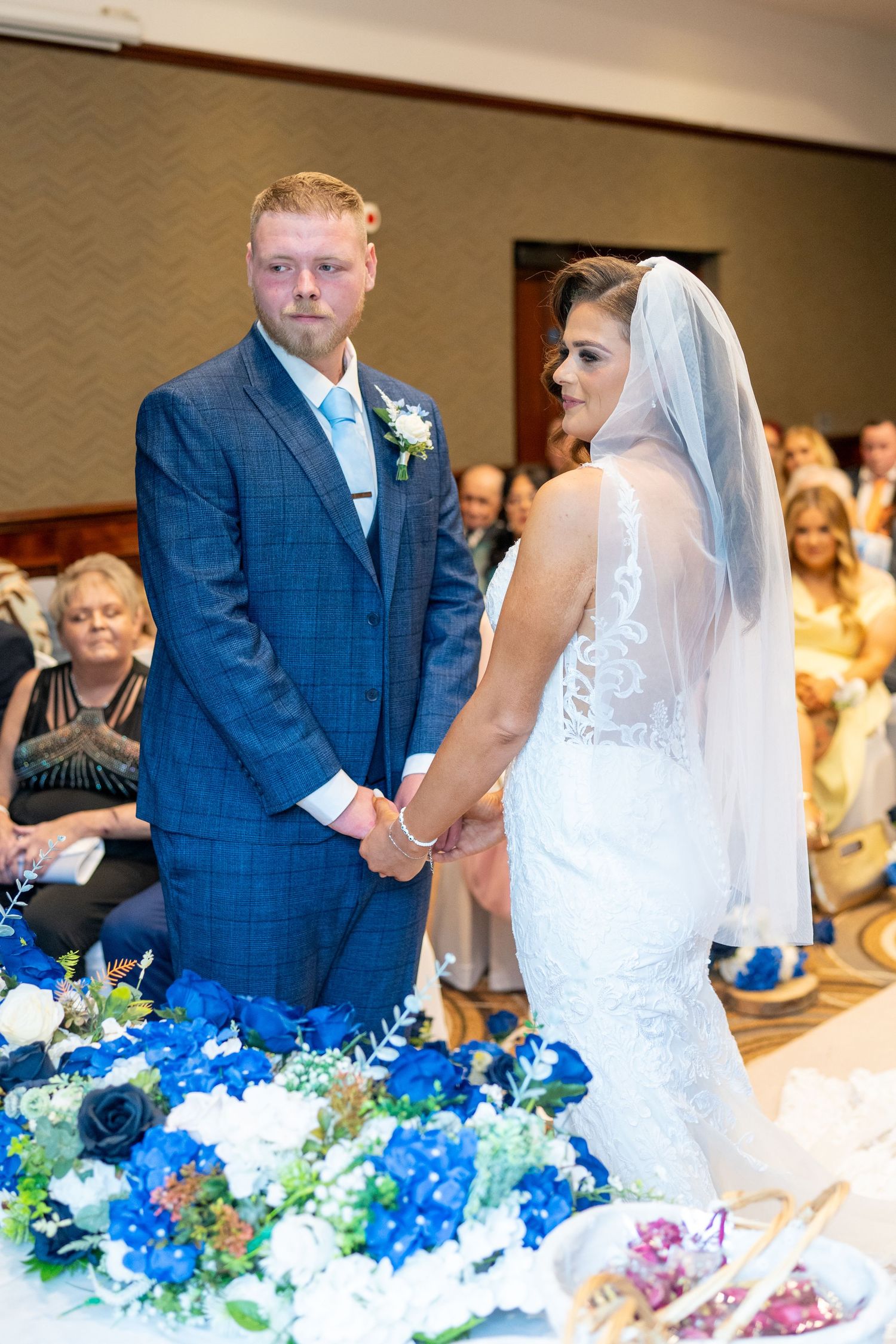 Bride and groom hold hands at floral-decorated altar during indoor wedding ceremony. Dave Martin Photographer Northern Ireland