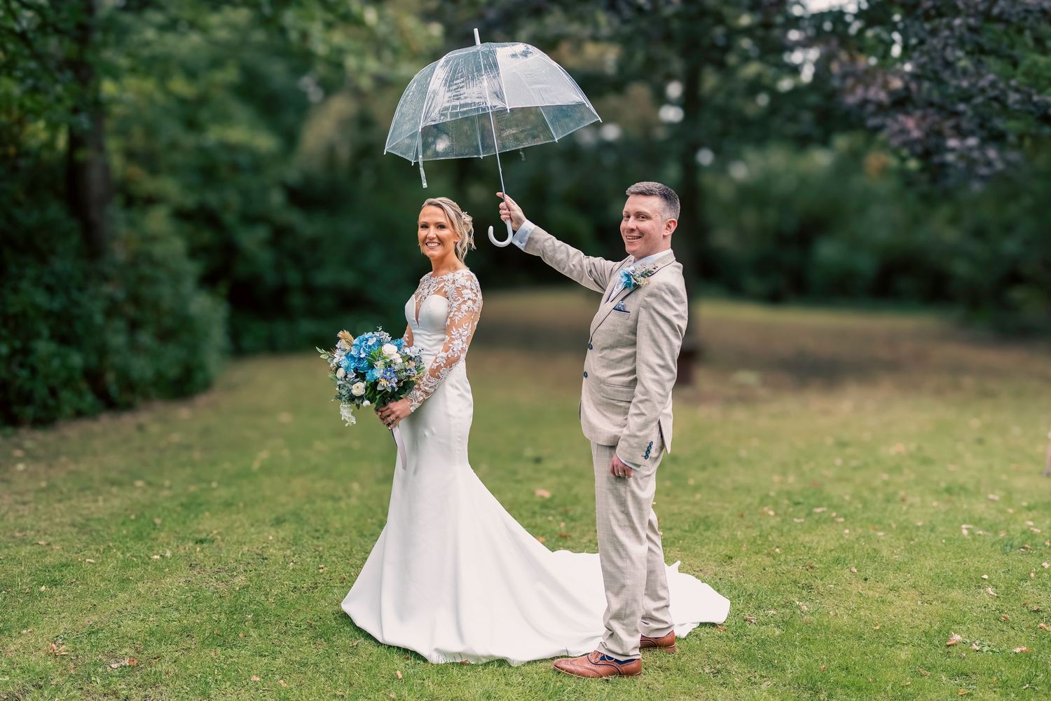 Bride in white gown holding bouquet stands with groom in tan suit holding clear umbrella on lush green lawn.