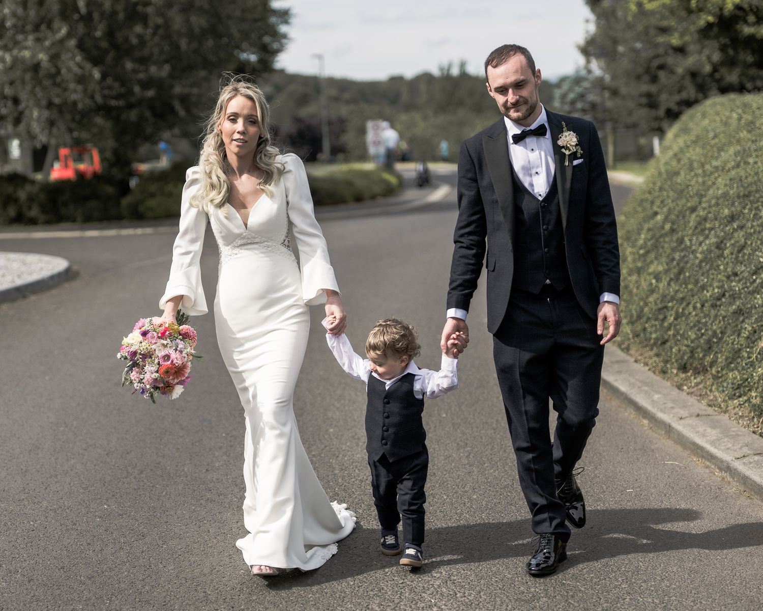 Bride in white gown and groom in black tuxedo walk with young boy on a sunny road, holding hands. ni wedding photographer Dave Martin