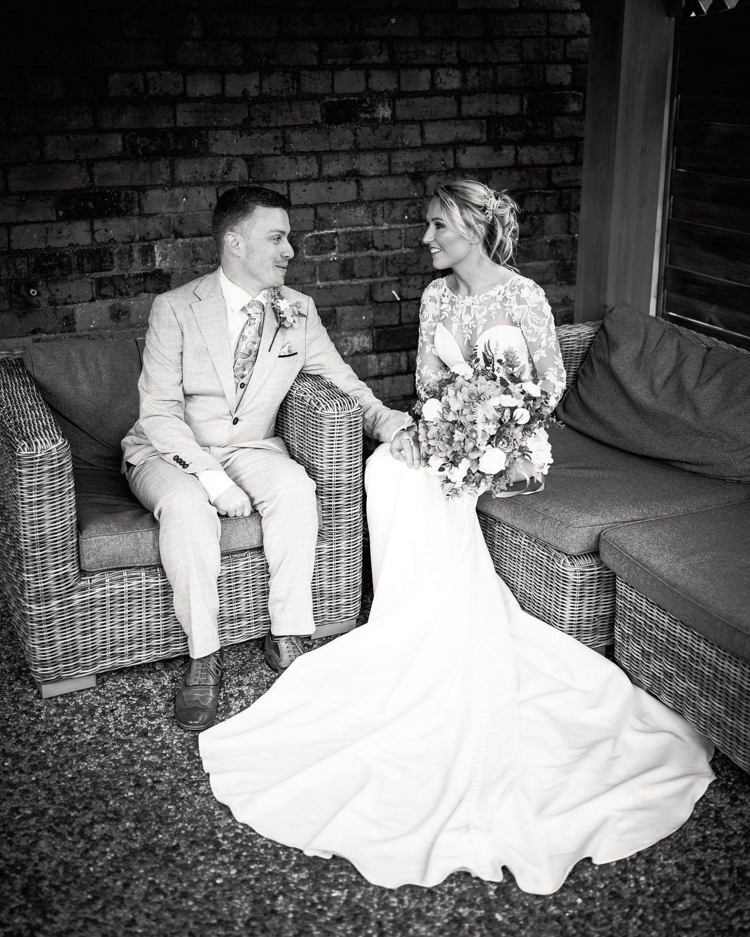 Black and white photo of a bride and groom sitting on wicker furniture, holding hands and smiling at each other on their wedding day.