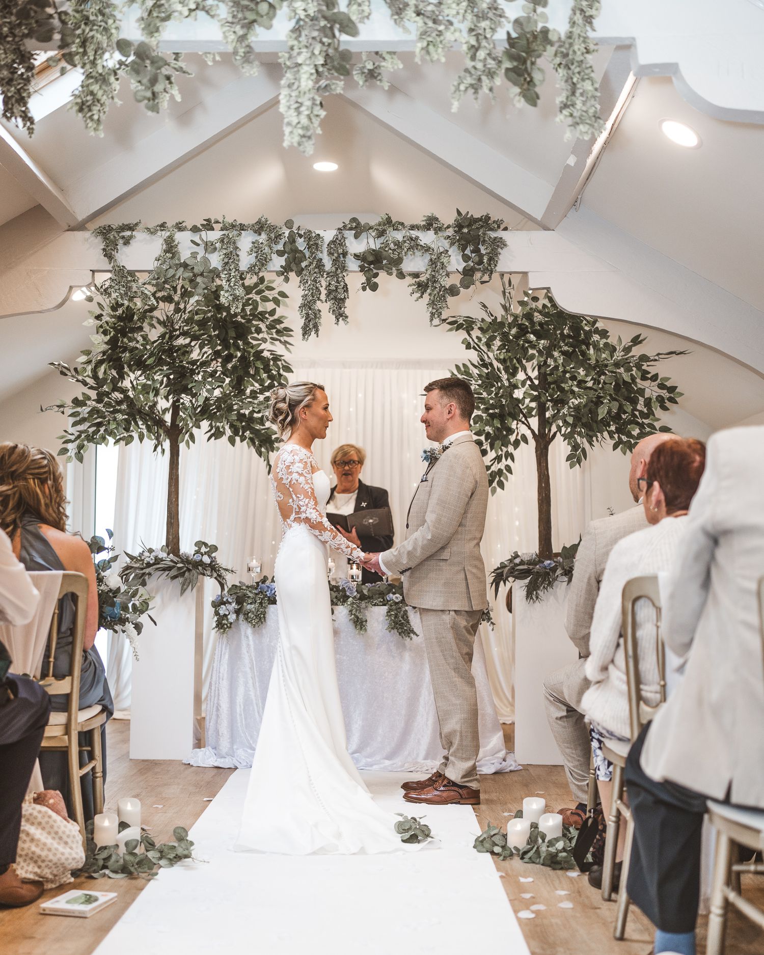 Bride and groom exchange vows at elegant indoor wedding ceremony with lush greenery trees and floral arch decor.