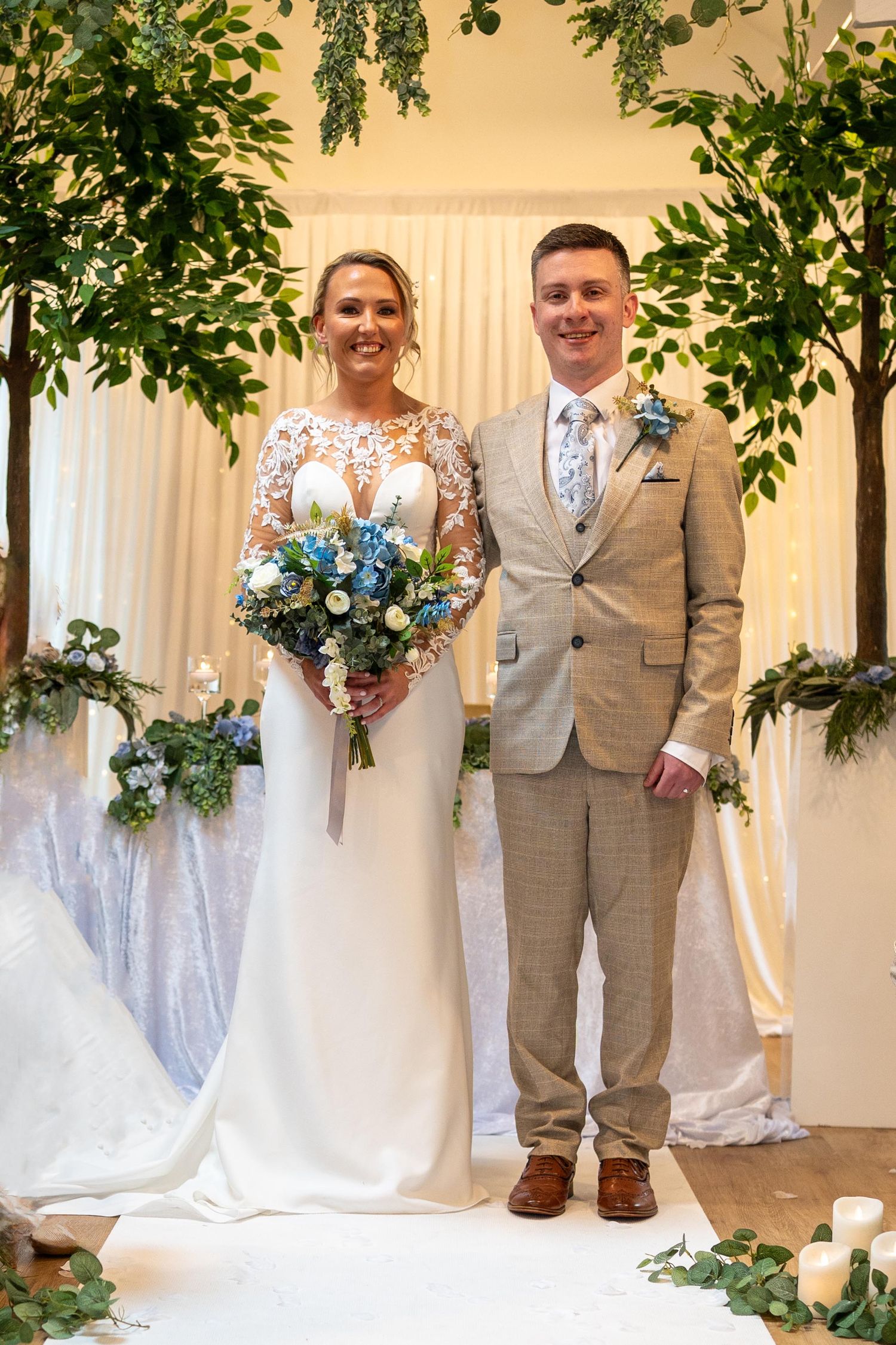 Bride in lace wedding gown holding blue bouquet stands with groom in tan suit before greenery arch.