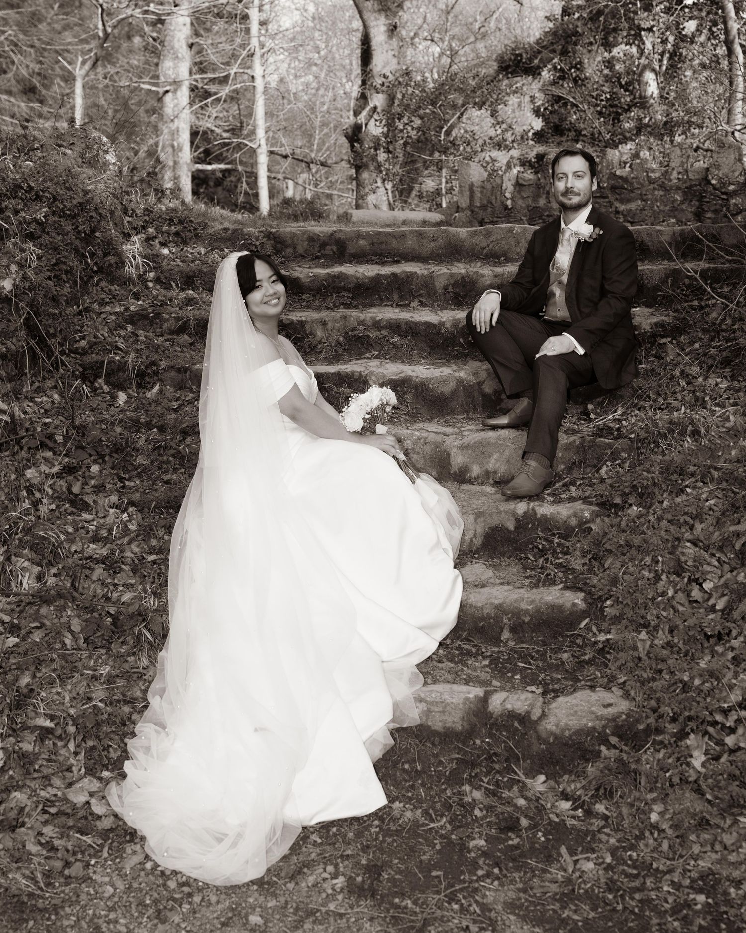 A bride in a white gown and veil sits on stone steps outdoors while the groom relaxes behind her in this sepia wedding photo. Dave Martin