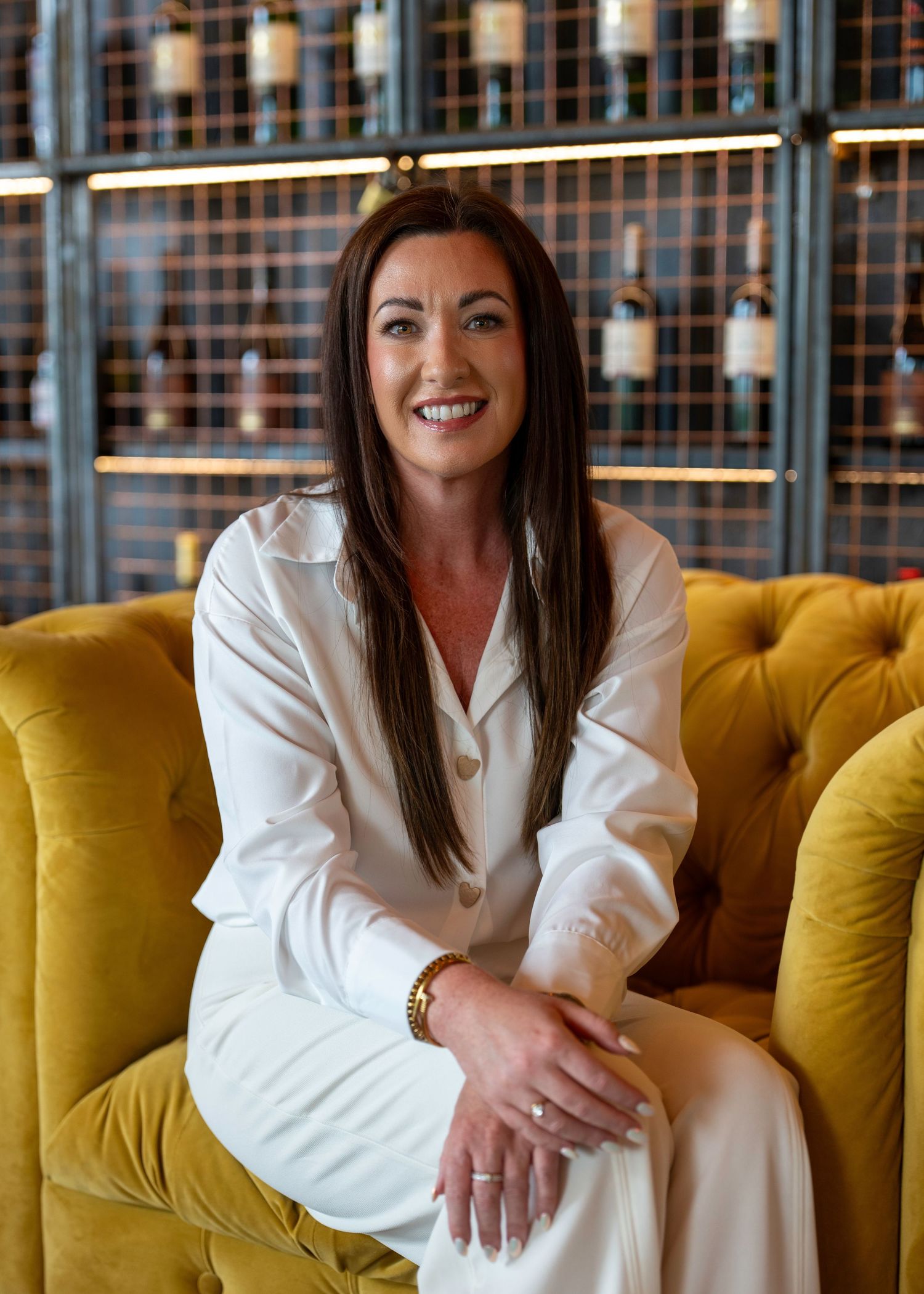 Smiling woman with dark hair in white outfit sitting on a gold velvet sofa in a restaurant wine bar setting. Dave Martin Photography