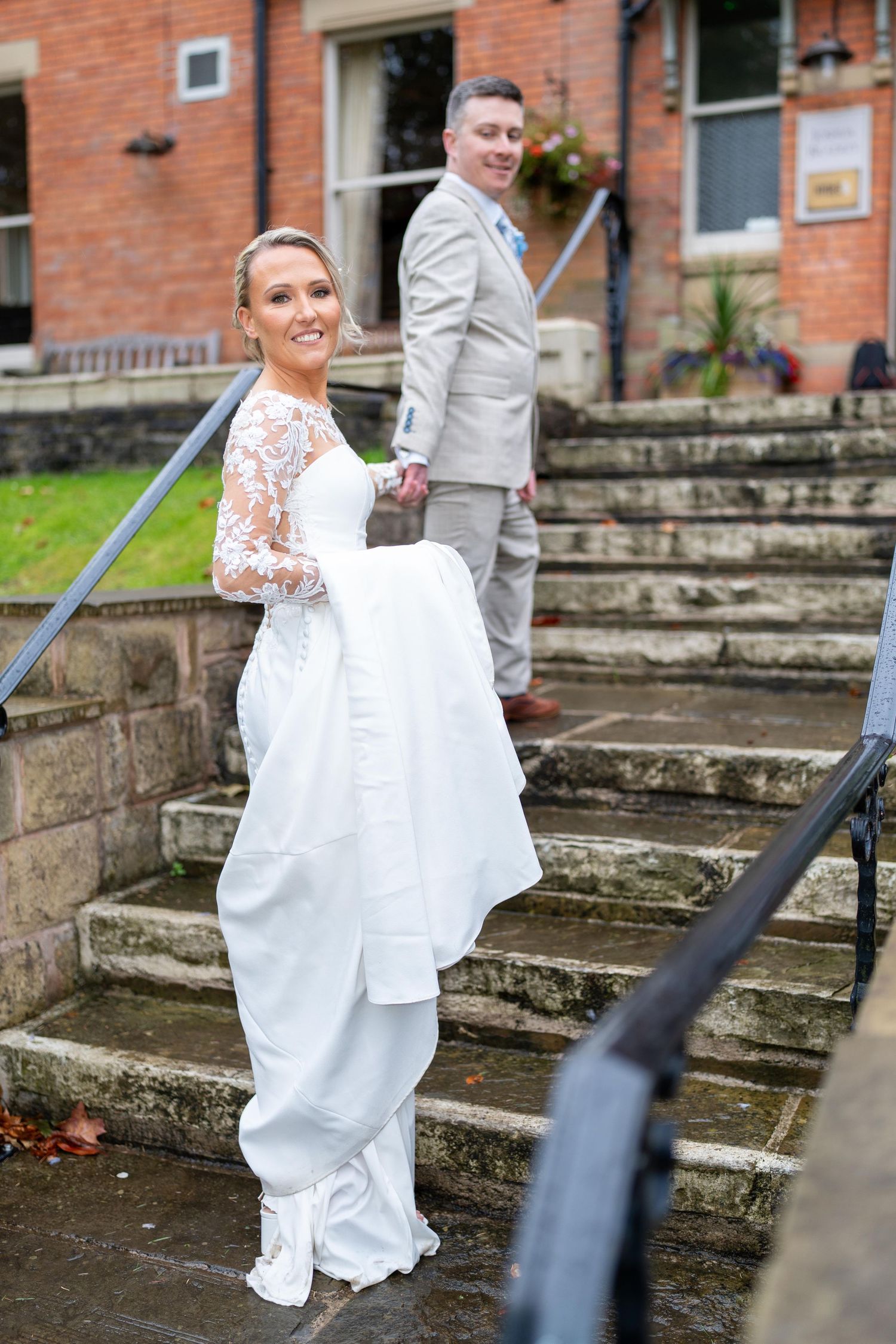 Bride in white lace gown holds dress on stone steps, smiling, with groom in grey suit behind her.