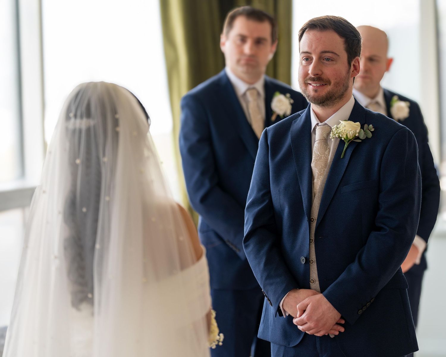 Groom in navy suit smiling at bride in white veil during wedding ceremony with groomsmen standing in background. Dave Martin Weddings