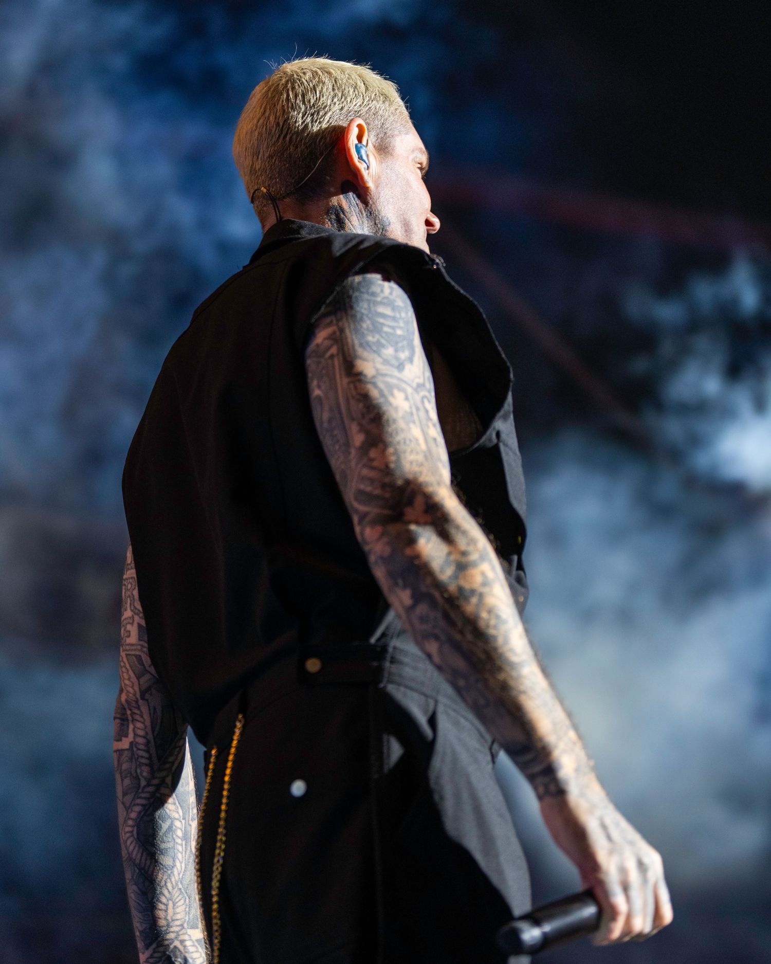 Shane Lynch with blonde hair in black vest viewed from behind holding microphone on smoky stage. Dave Martin Photography