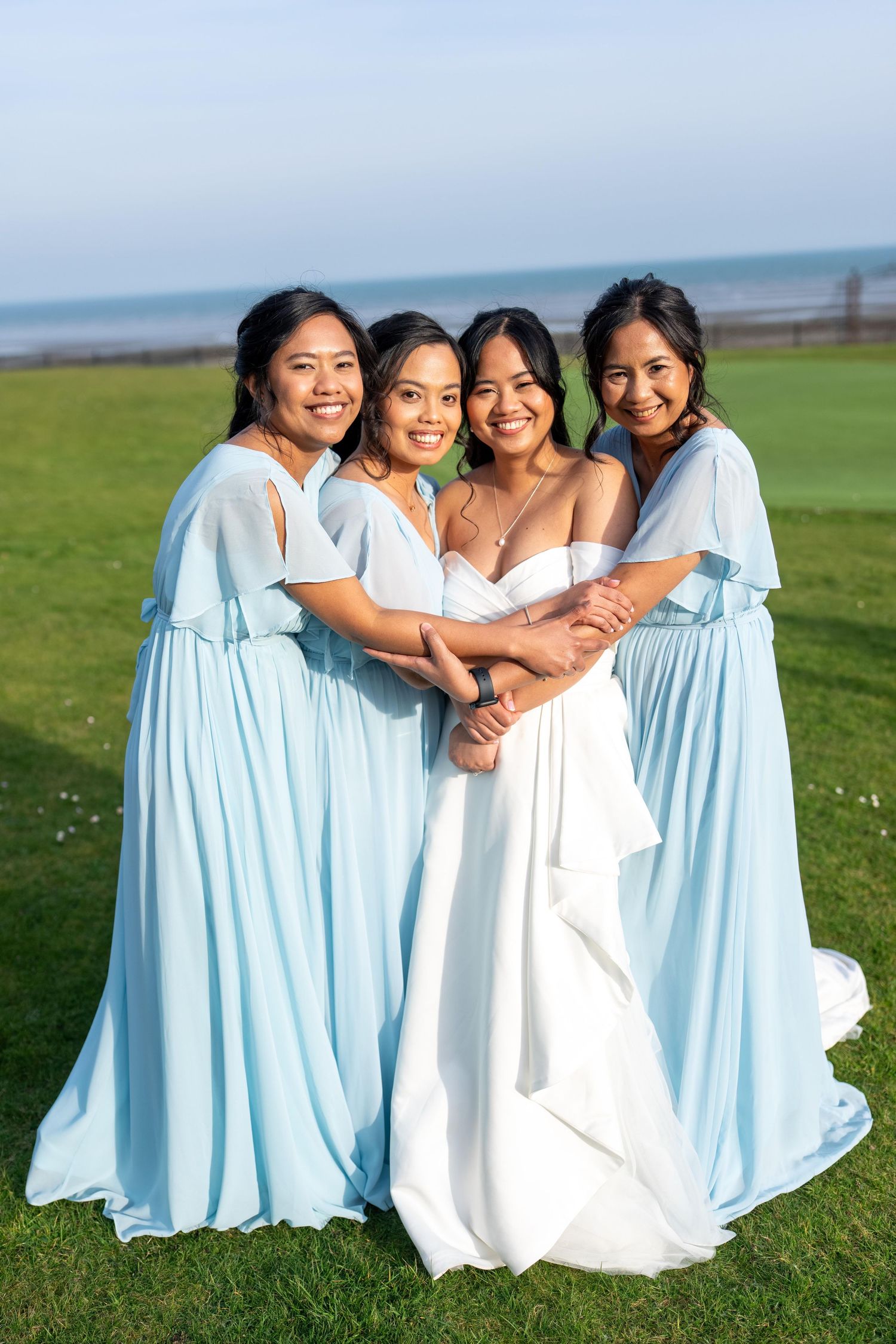 Bride in white gown poses with three bridesmaids in light blue dresses on green lawn with ocean backdrop. Dave Martin Weddings