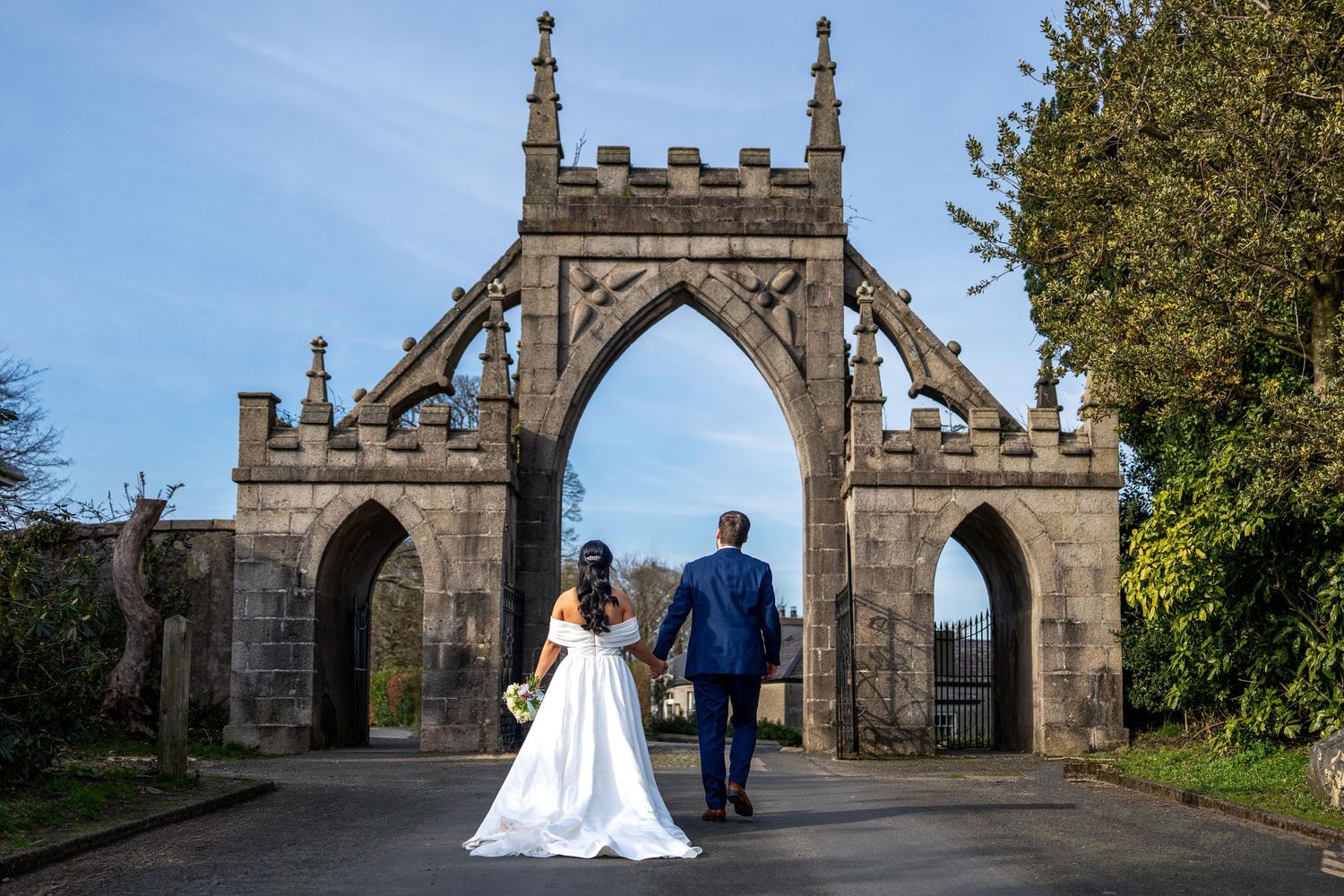 Bride and groom walking hand in hand toward a stunning Gothic stone archway at an elegant outdoor wedding venue.