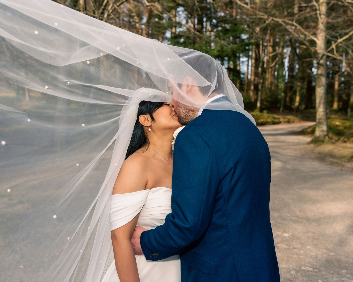 Bride and groom share a romantic kiss under a flowing white veil outdoors, surrounded by trees. By Dave Martin Photography