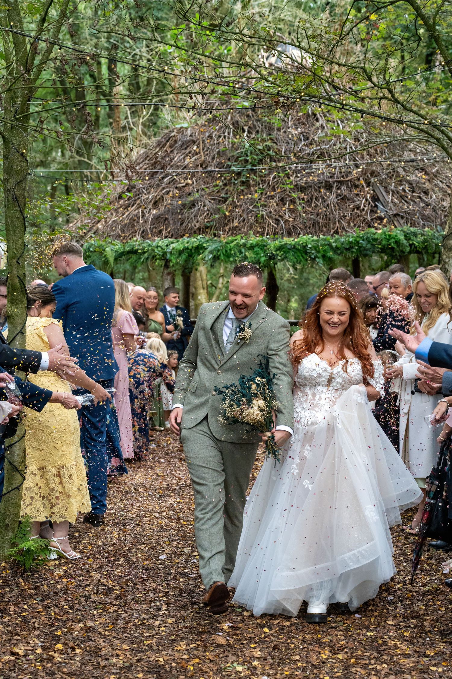 Newlywed couple walks through confetti-throwing guests along woodland path after outdoor wedding ceremony. Dave Martin Photographer Northern