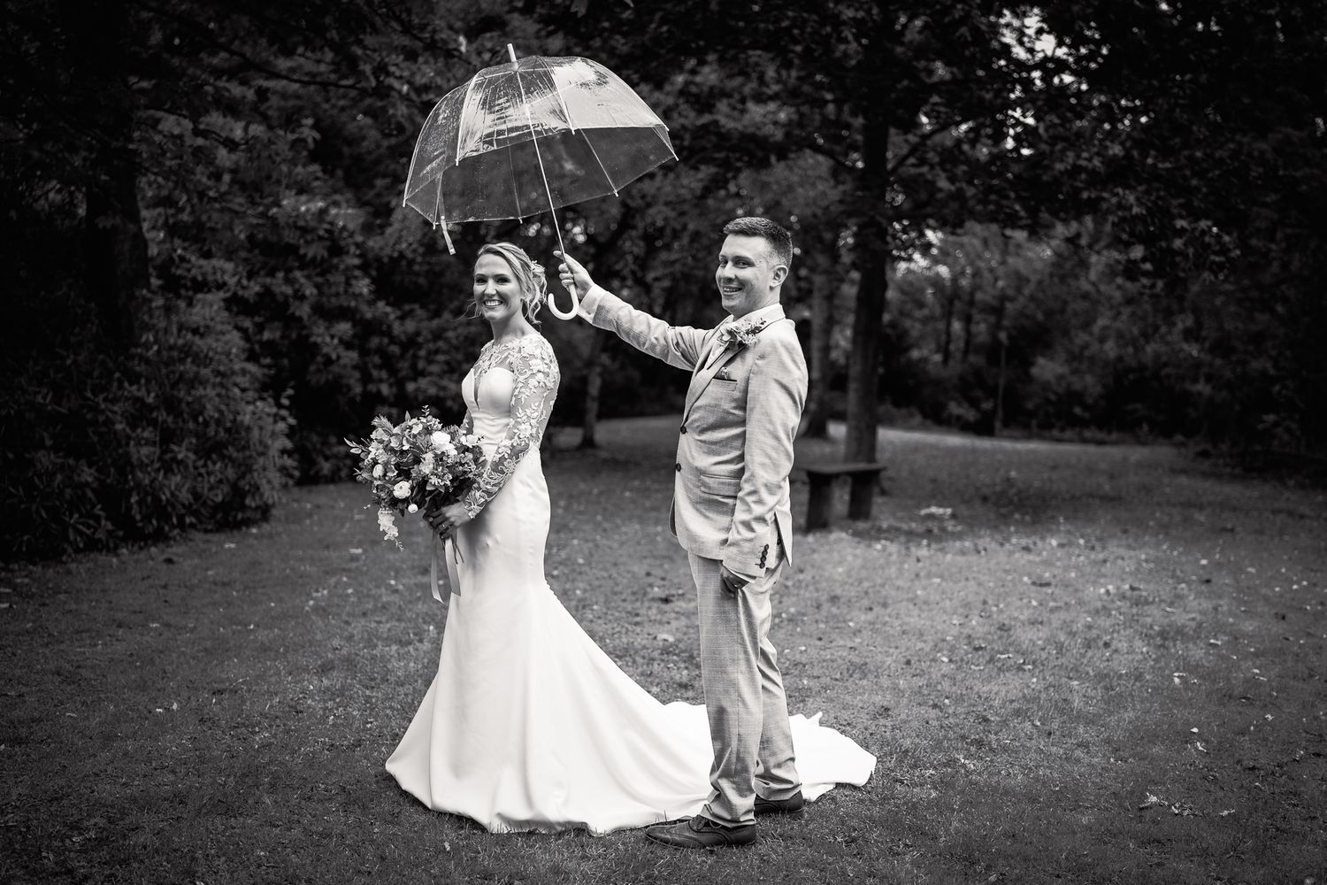 Black and white photo of a bride and groom posing outdoors under a clear umbrella on their rainy wedding day. Dave Martin wedding Photos