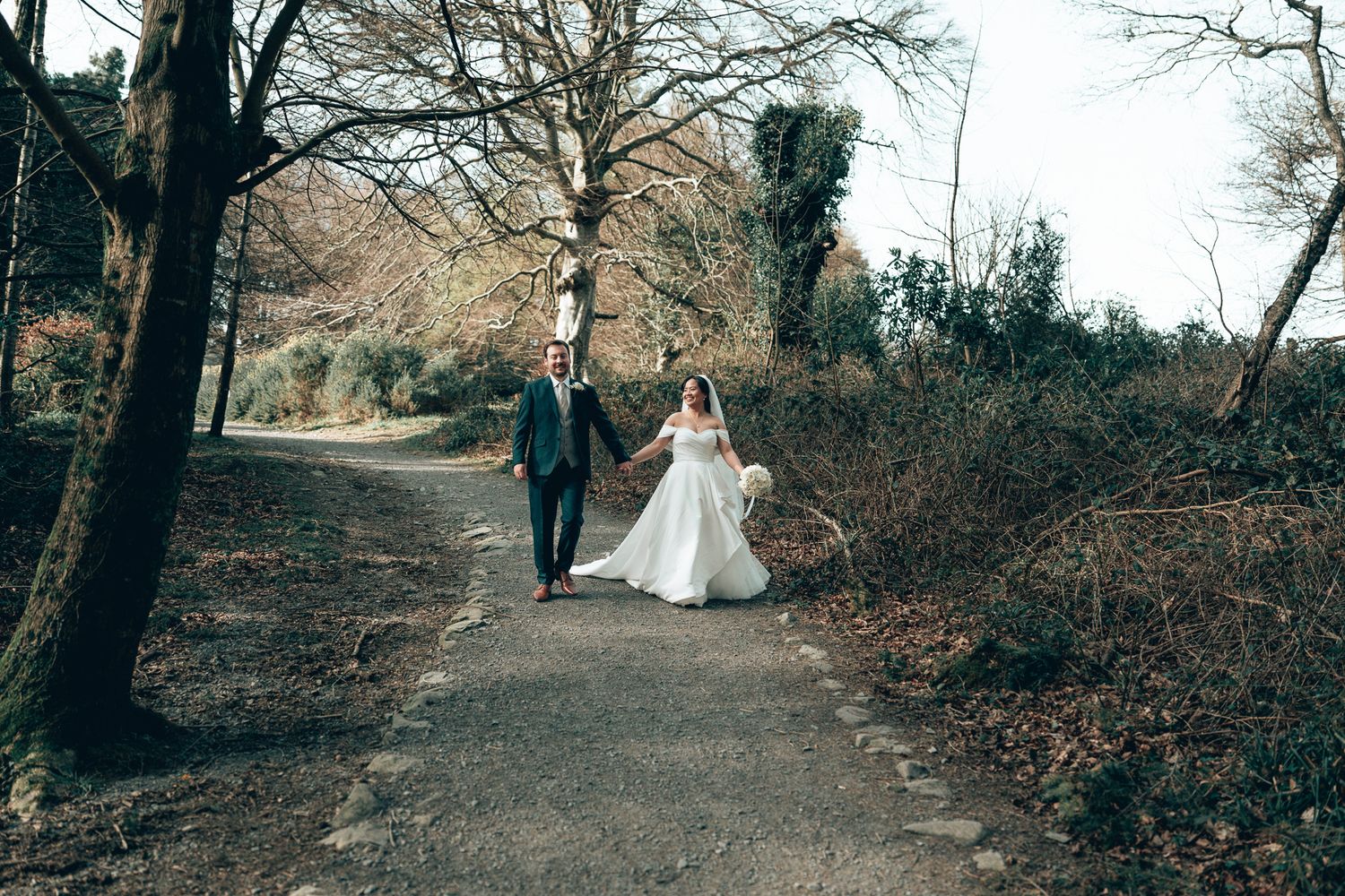 Bride and groom walking hand in hand along a scenic woodland path on their wedding day. Dave Martin wedding Photographer NI
