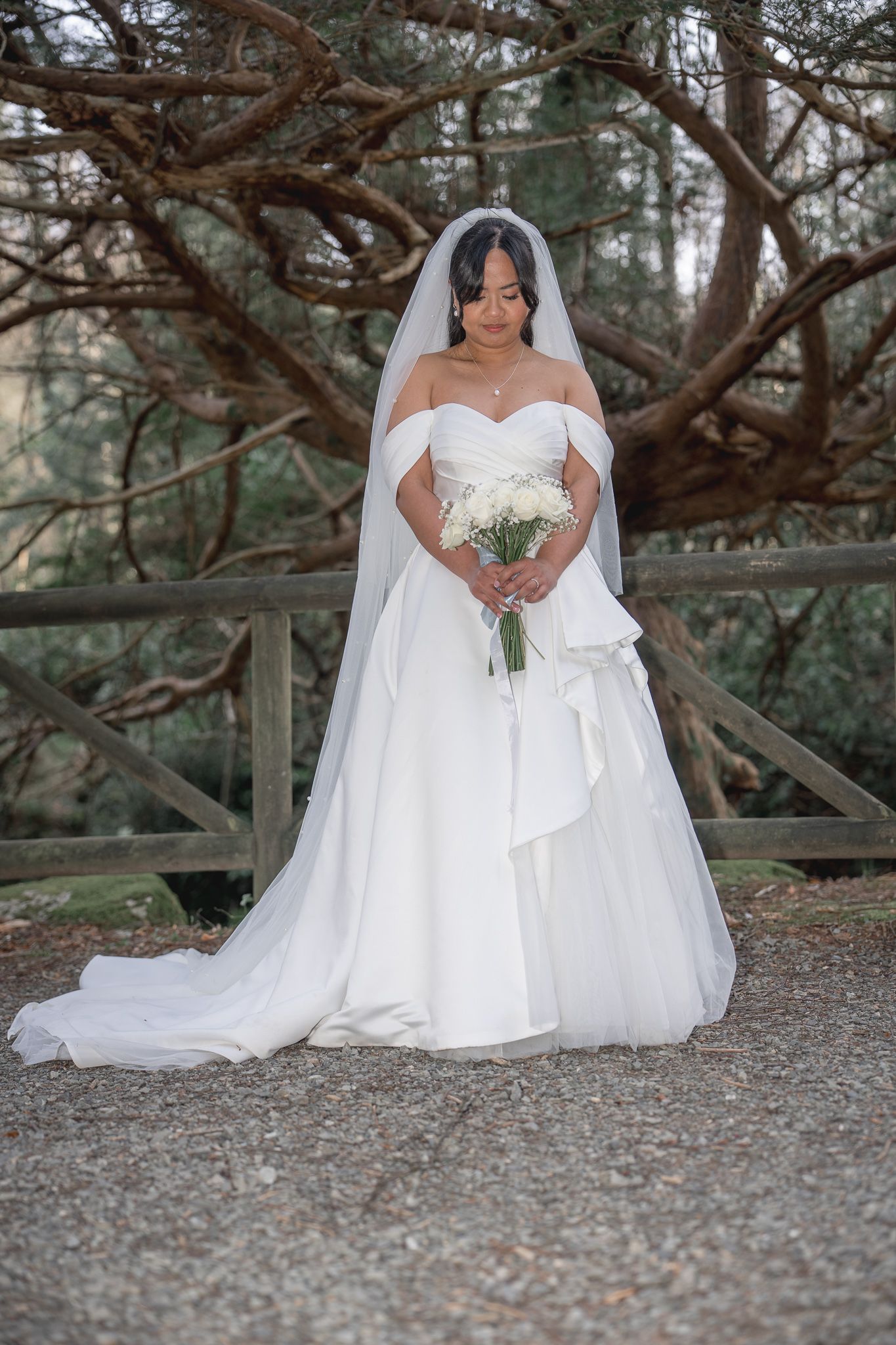 Bride in white off-shoulder gown with veil holds bouquet outdoors near wooden fence and trees. Dave Martin wedding Photographer NI