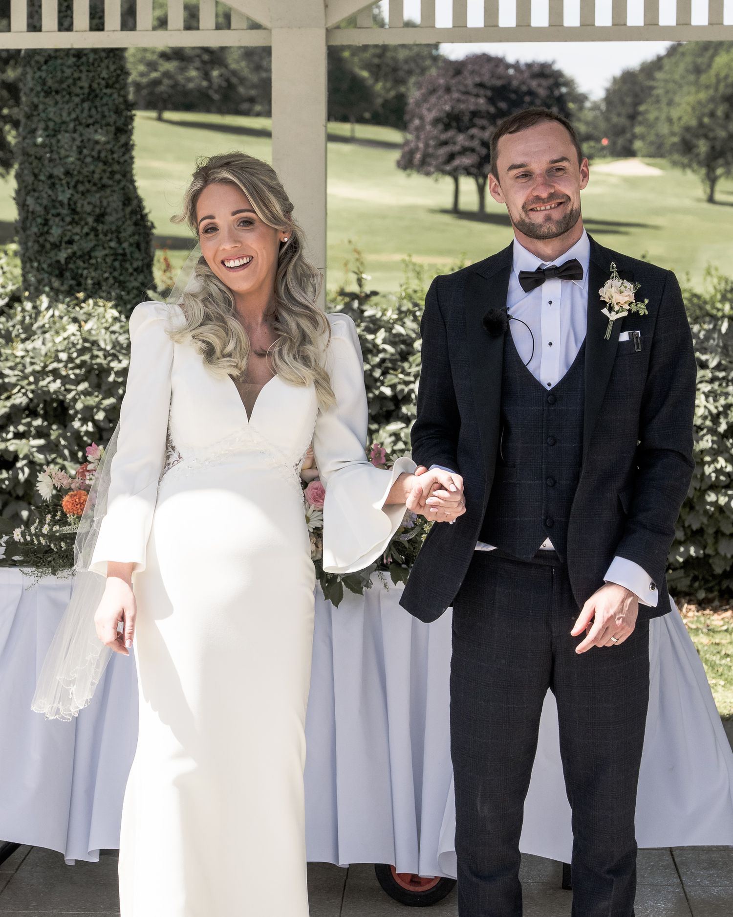 A bride in a white long-sleeve gown and groom in a black tuxedo hold hands and smile at an outdoor wedding ceremony. Dave Martin wedding
