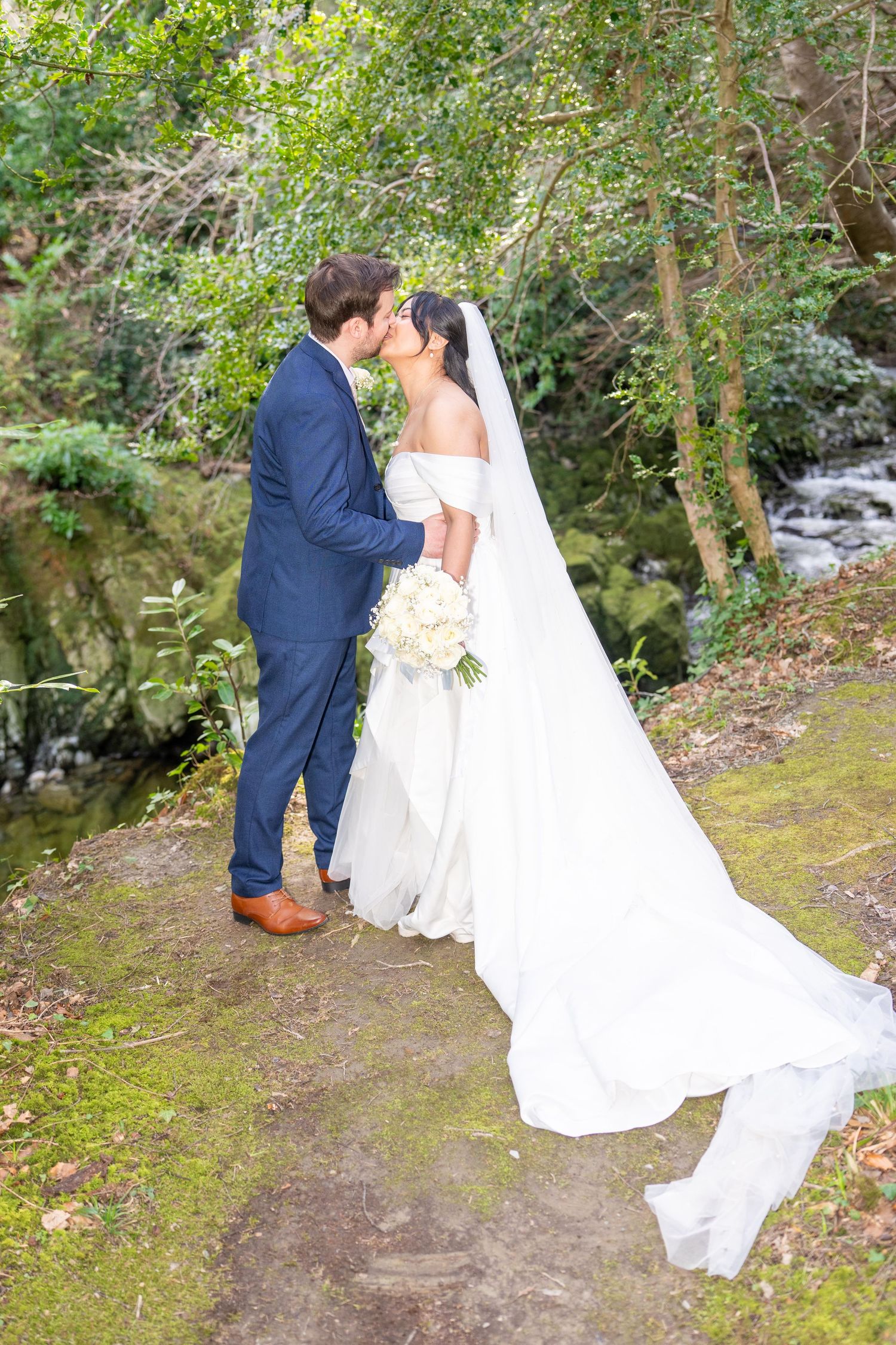 Bride and groom share a romantic kiss outdoors near a stream, she in a white gown with veil, he in a blue suit. Tollymore forest