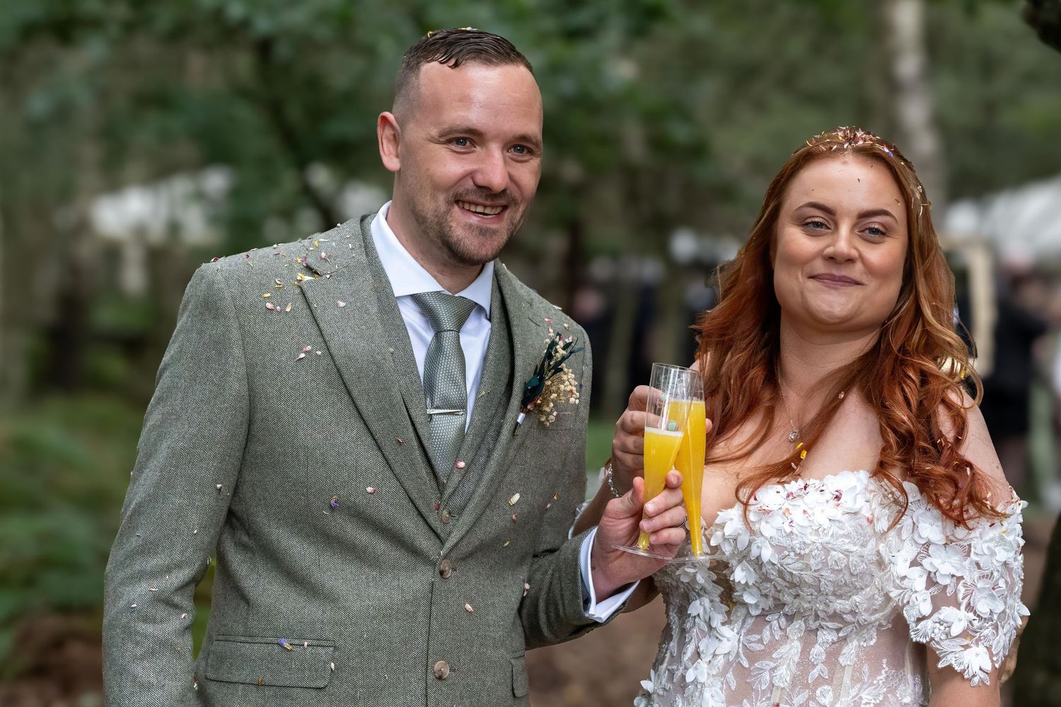 Bride and groom toasting with champagne flutes outdoors, she in lace gown, he in grey tweed suit.