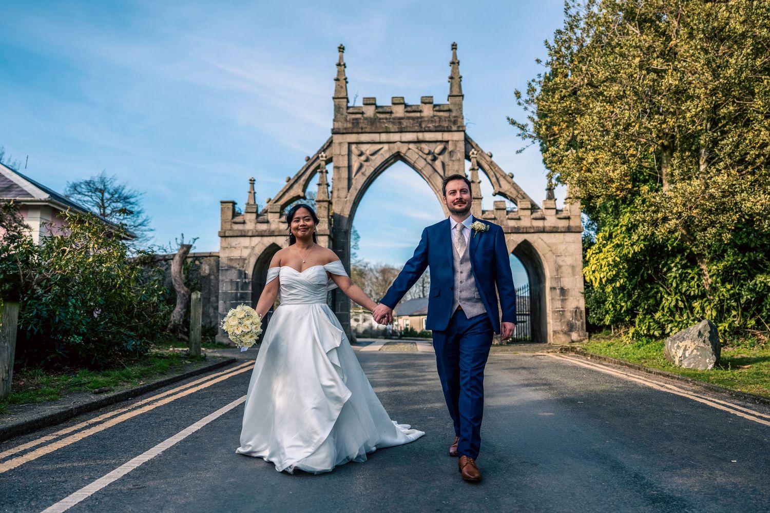 Bride and groom walking hand in hand through a stone Gothic arch gateway on their wedding day. By Dave Martin Photography