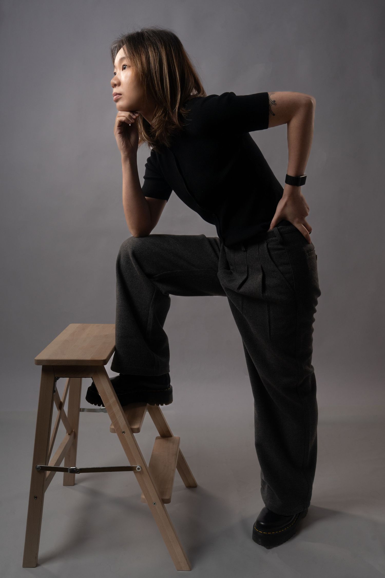 Young woman in black top and cargo pants poses confidently on wooden step stool against gray studio background. Dave Martin Photography