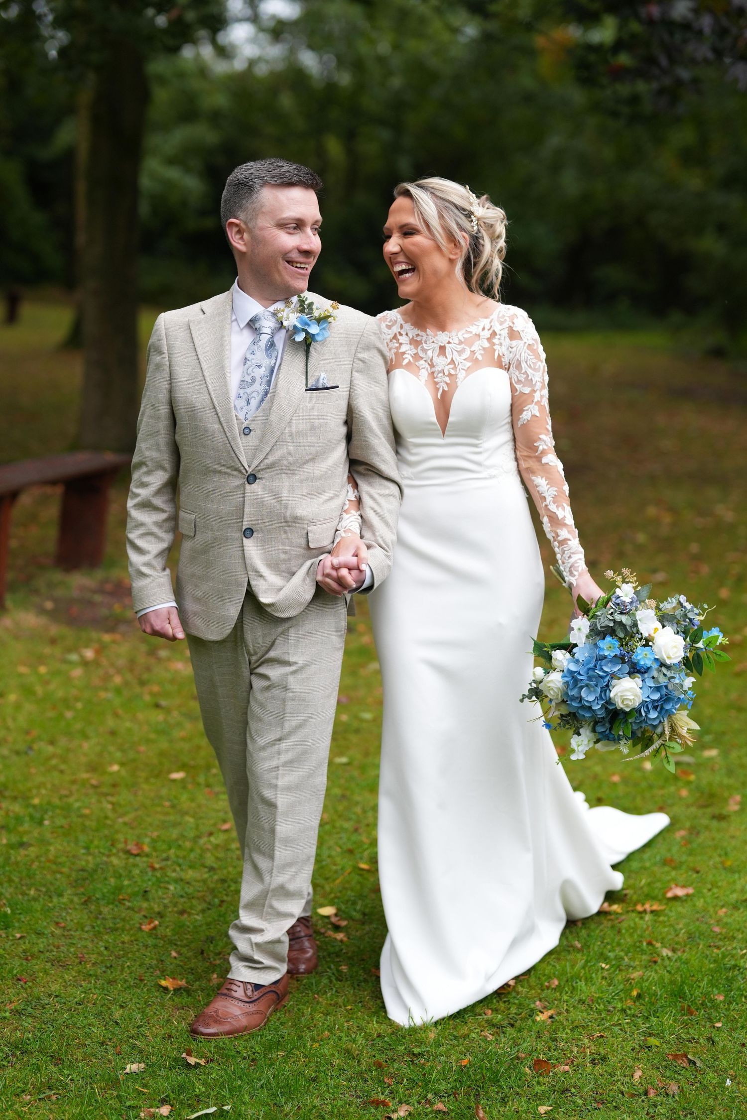 Laughing bride and groom walk hand in hand outdoors, she holds blue bouquet, he wears tan suit.