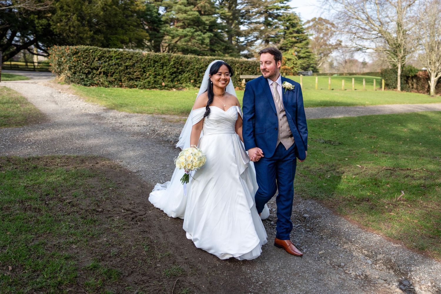 A bride and groom walk hand in hand along a garden path, she in a white gown with veil, he in a navy suit. Dave Martin Weddings