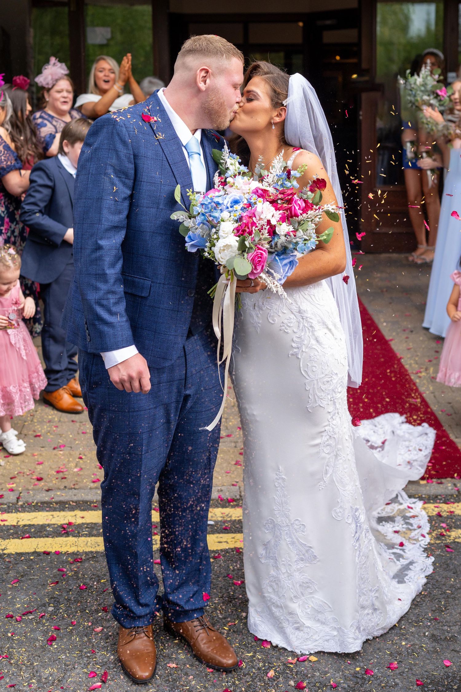 Bride and groom share a kiss outside wedding venue as guests throw colorful confetti, bride holds vibrant bouquet.