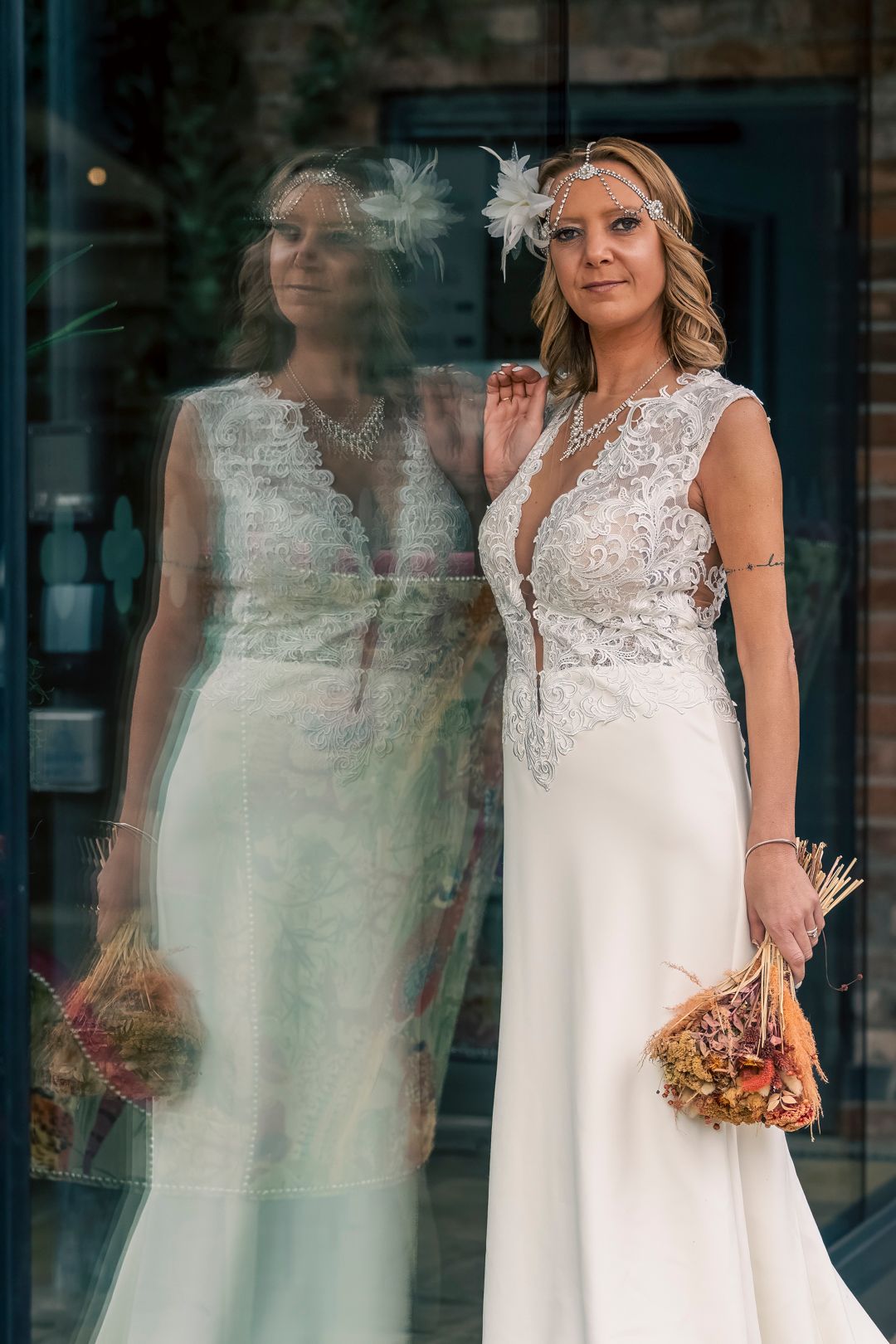 Bride in lace wedding dress holding bouquet, reflected in glass window, wearing floral headpiece. Dave Martin Photography