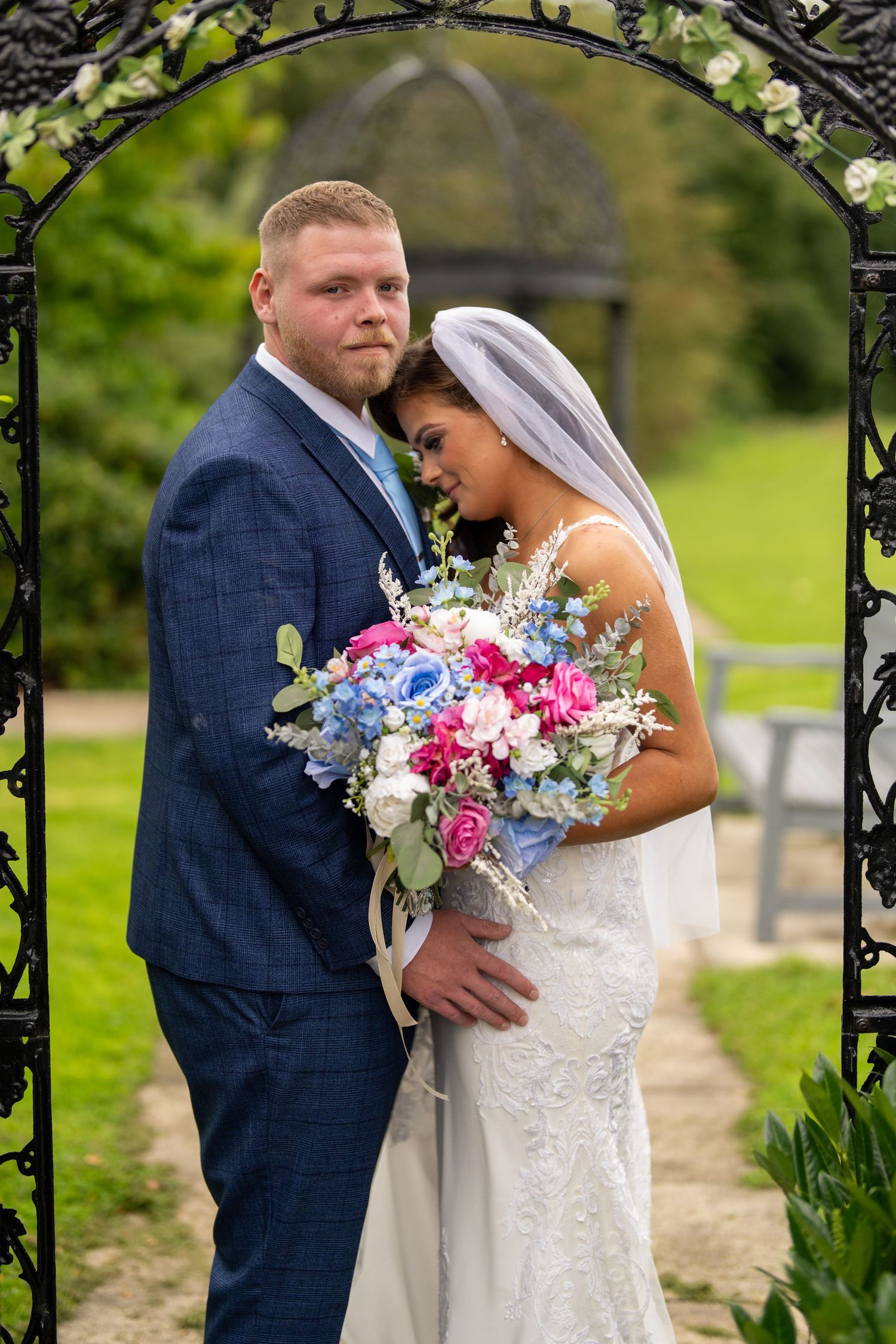Bride and groom posing under decorative arch, bride holding vibrant blue and pink floral bouquet. Dave Martin Photographer Northern Ireland