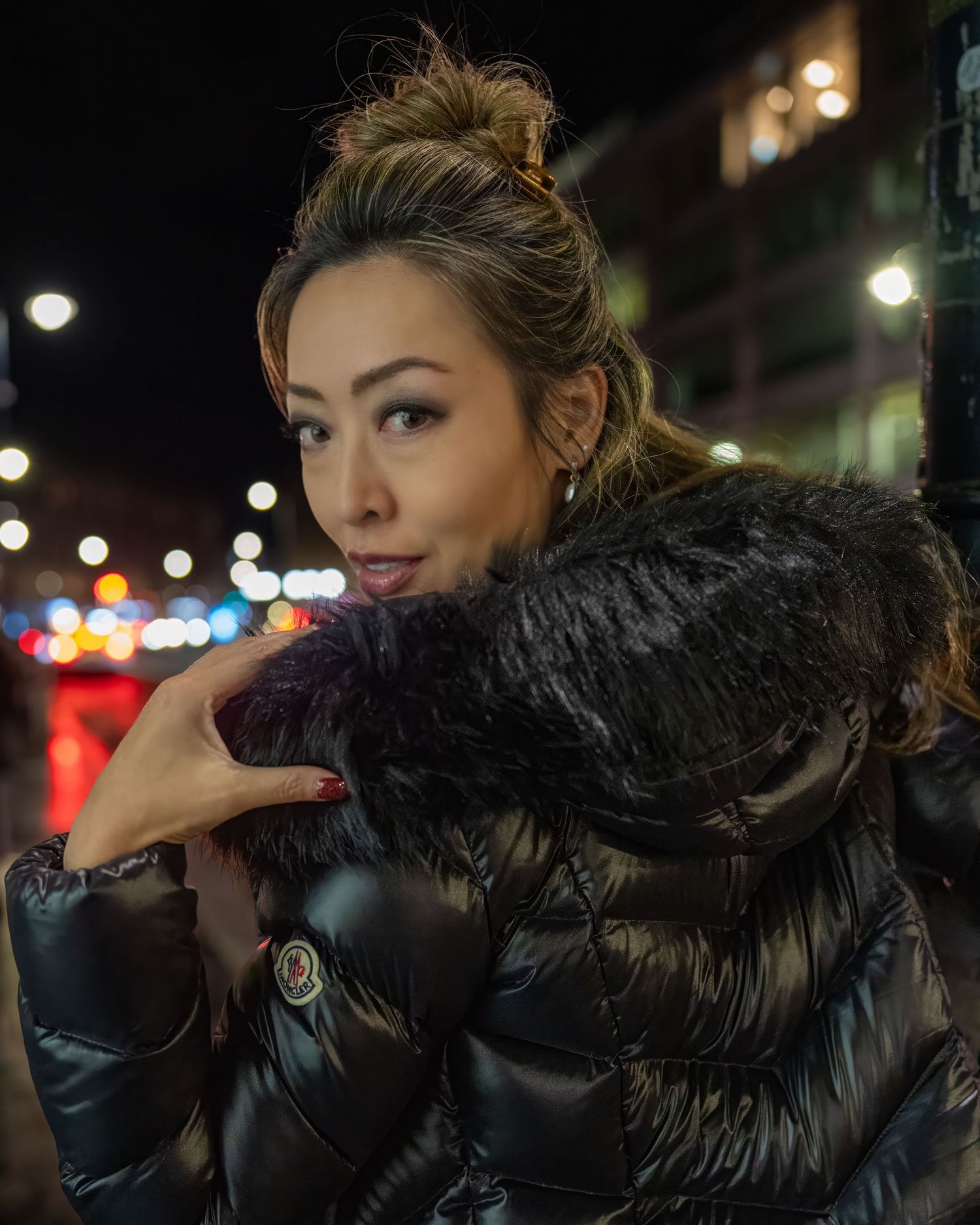 Woman in black fur-trimmed Moncler puffer jacket posing outdoors on city street at night. Dave Martin celebrity Photography