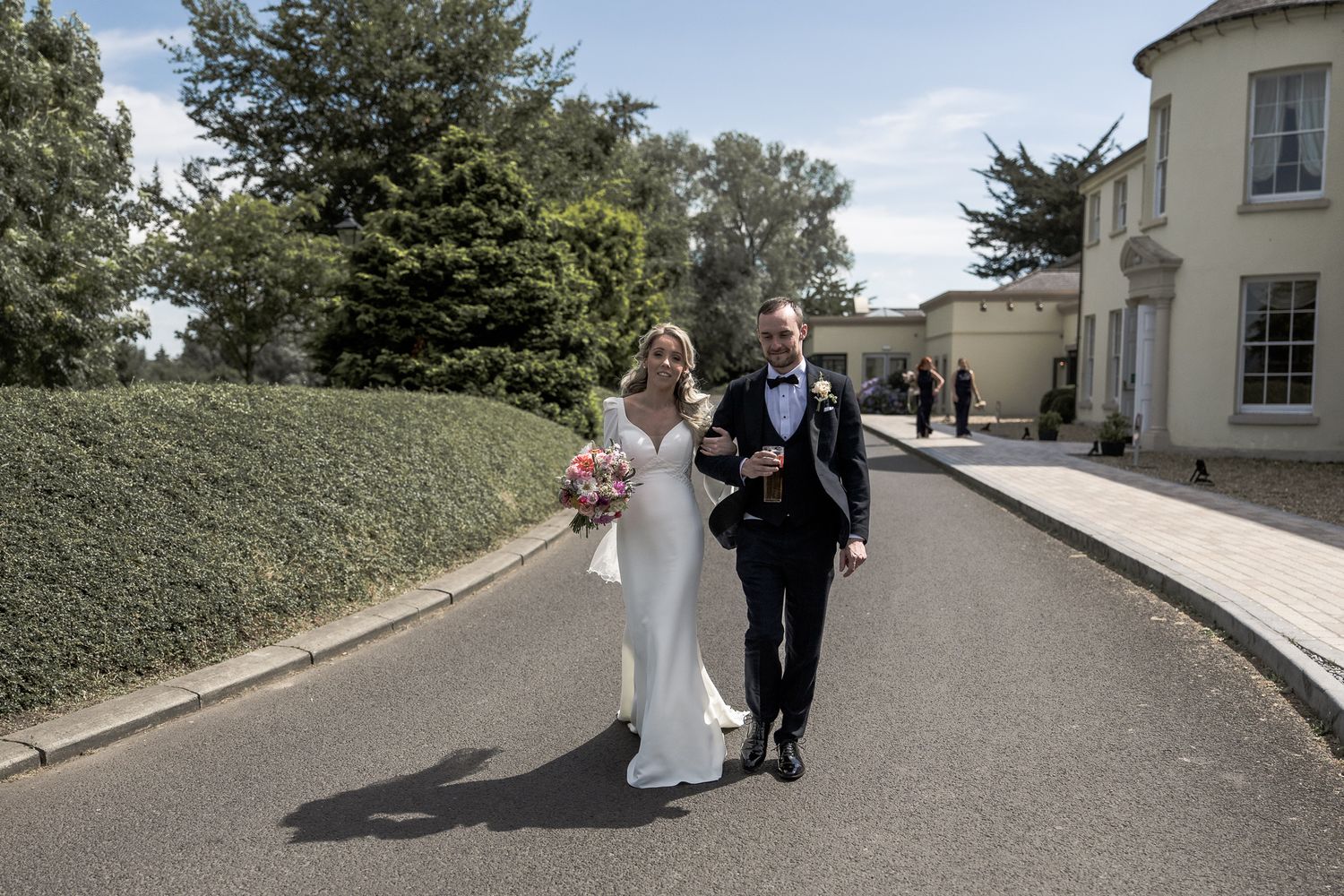 Bride and groom walking hand in hand down a tree-lined driveway outside an elegant manor wedding venue. Northern ireland weddings