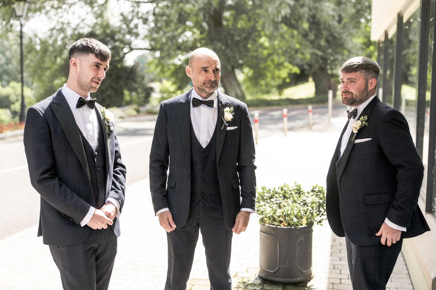 Three groomsmen in black tuxedos with boutonnieres conversing outdoors on a sunny wedding day. Dave Martin wedding Photographer NI