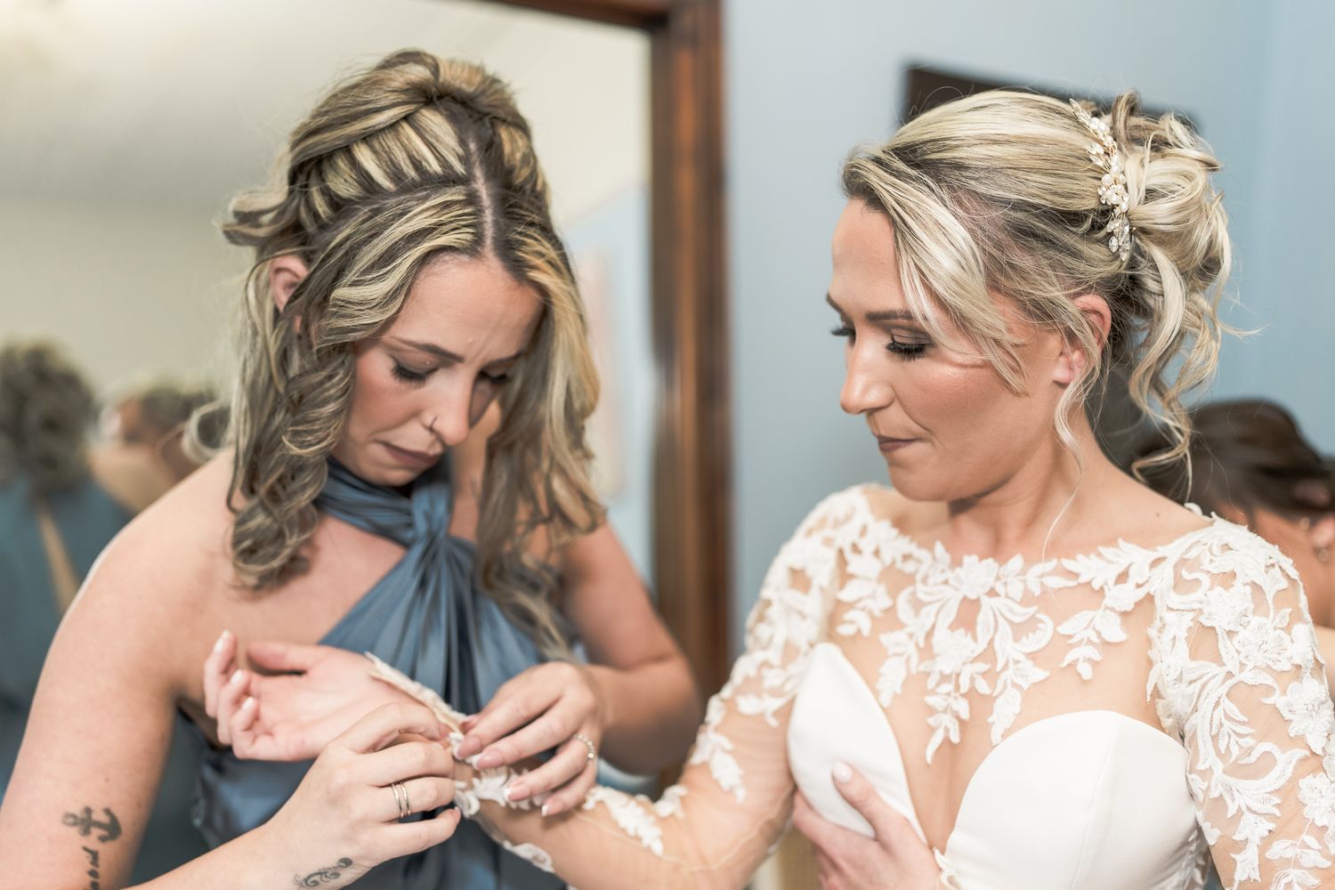 Bride in lace gown gets bracelet help from bridesmaid in blue dress during wedding preparations. Dave Martin wedding Photographer NI