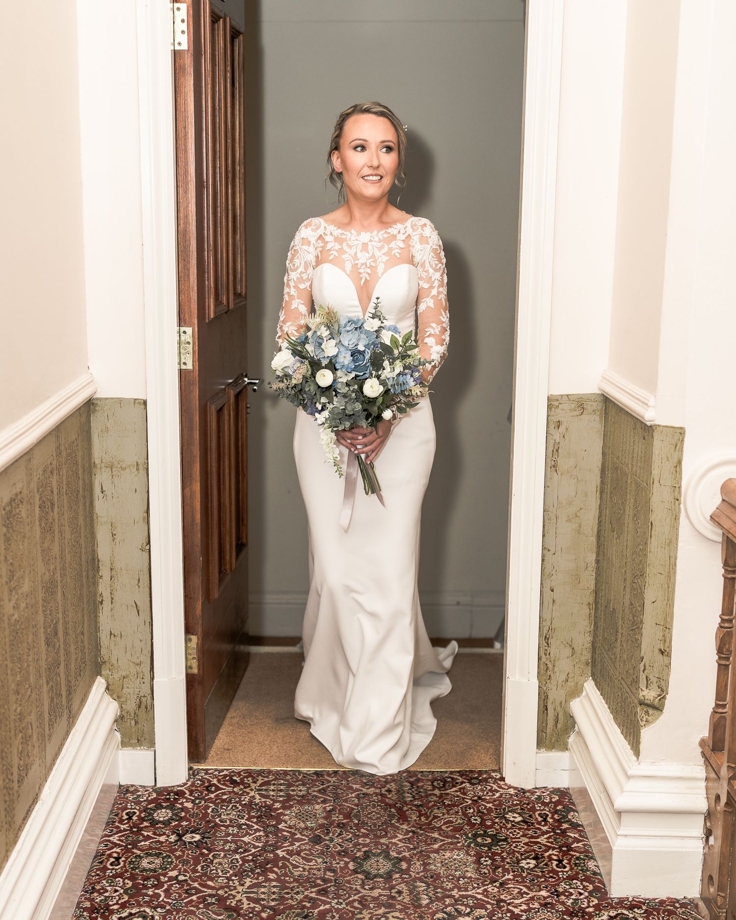 Bride in lace long-sleeve gown holding blue and white bouquet stands in doorway of elegant venue. Dave Martin wedding Photographer NI