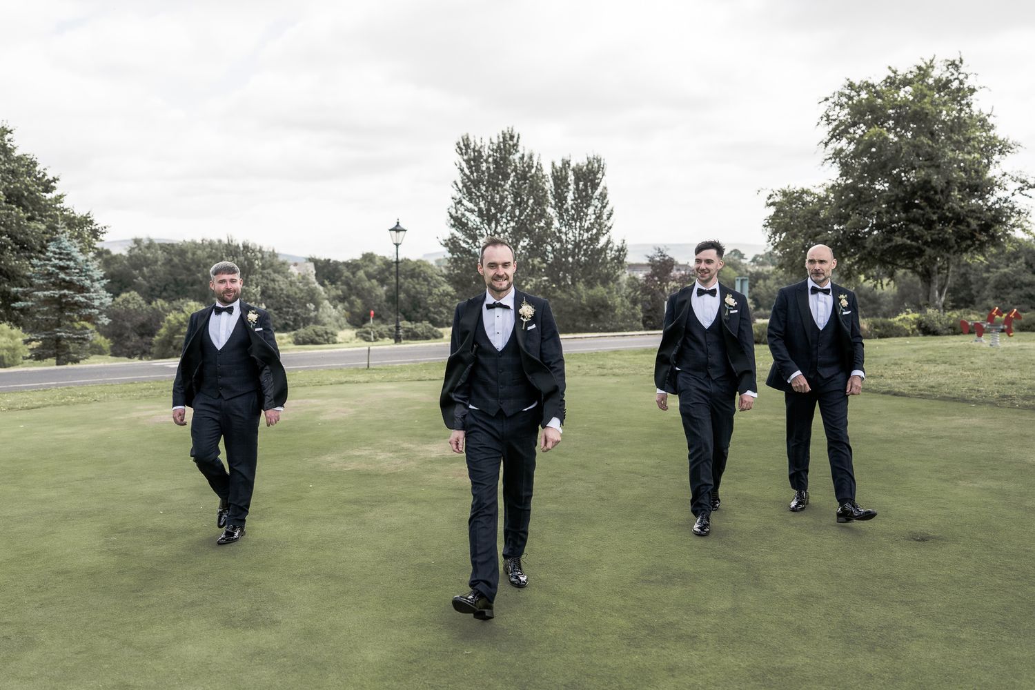 Four groomsmen in black tuxedos walking confidently across a green lawn at an outdoor wedding venue. Dave Martin wedding Photographer NI
