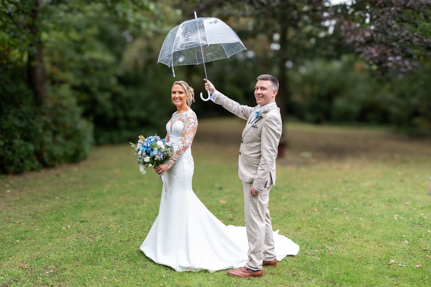 Groom holds clear umbrella over bride in lace wedding gown on green lawn in rain. Dave Martin Photographer Northern Ireland