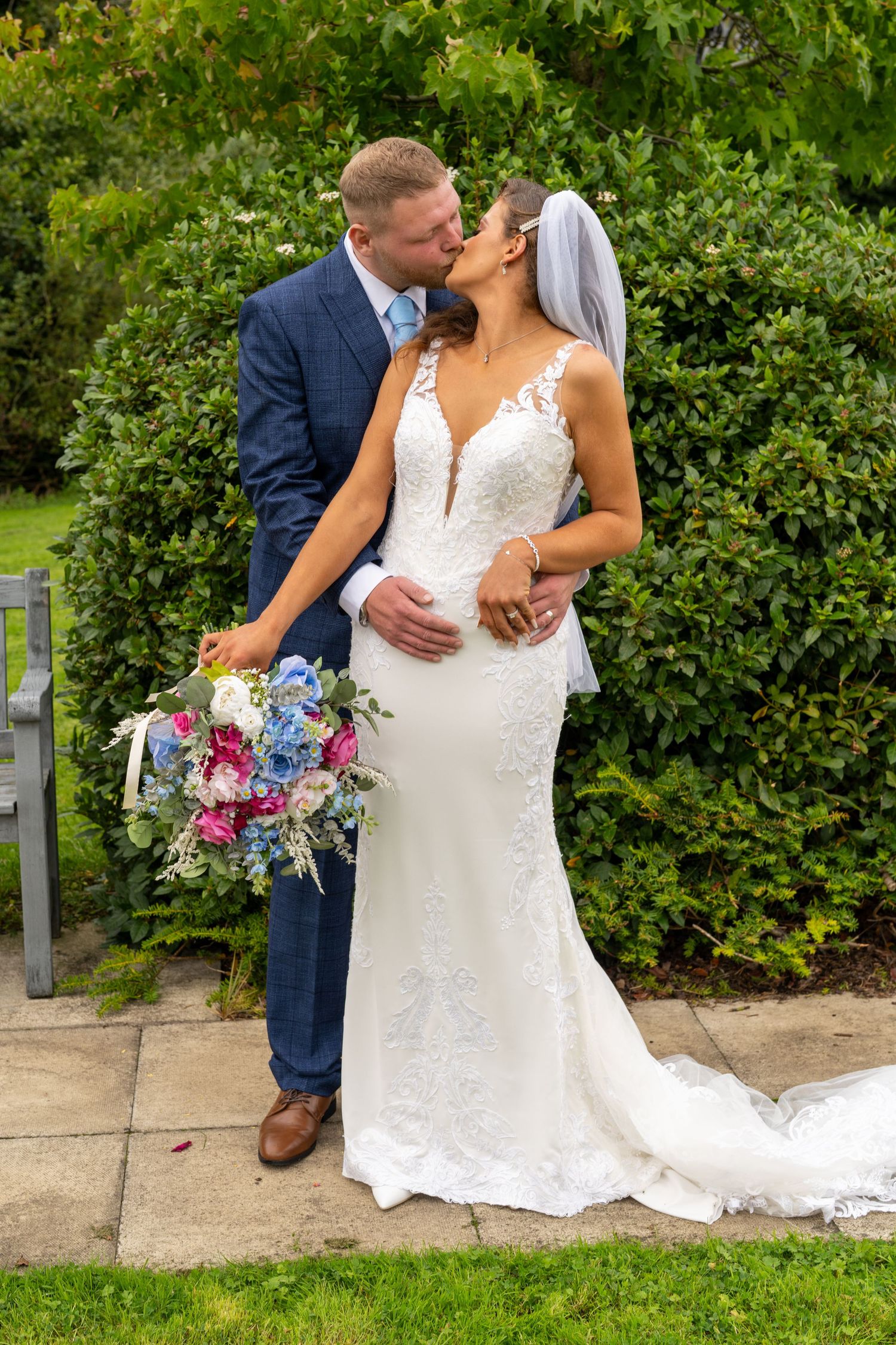 Bride and groom sharing a kiss in garden, bride in fitted lace gown holding colorful bouquet. Dave Martin Photographer Northern Ireland