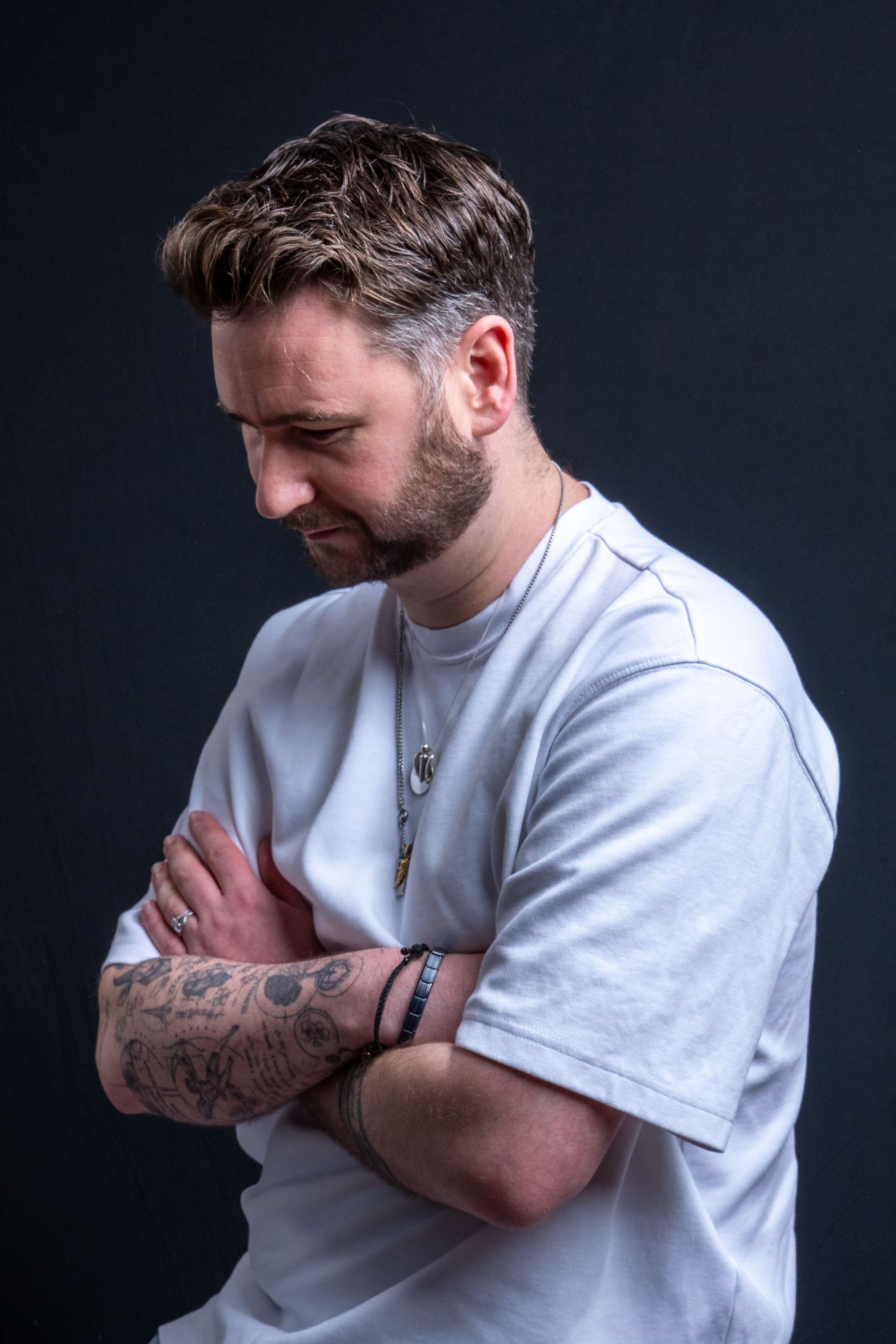 A tattooed bearded man in a white t-shirt with arms crossed looking down on dark background. Dave Martin Photography Studio Belfast