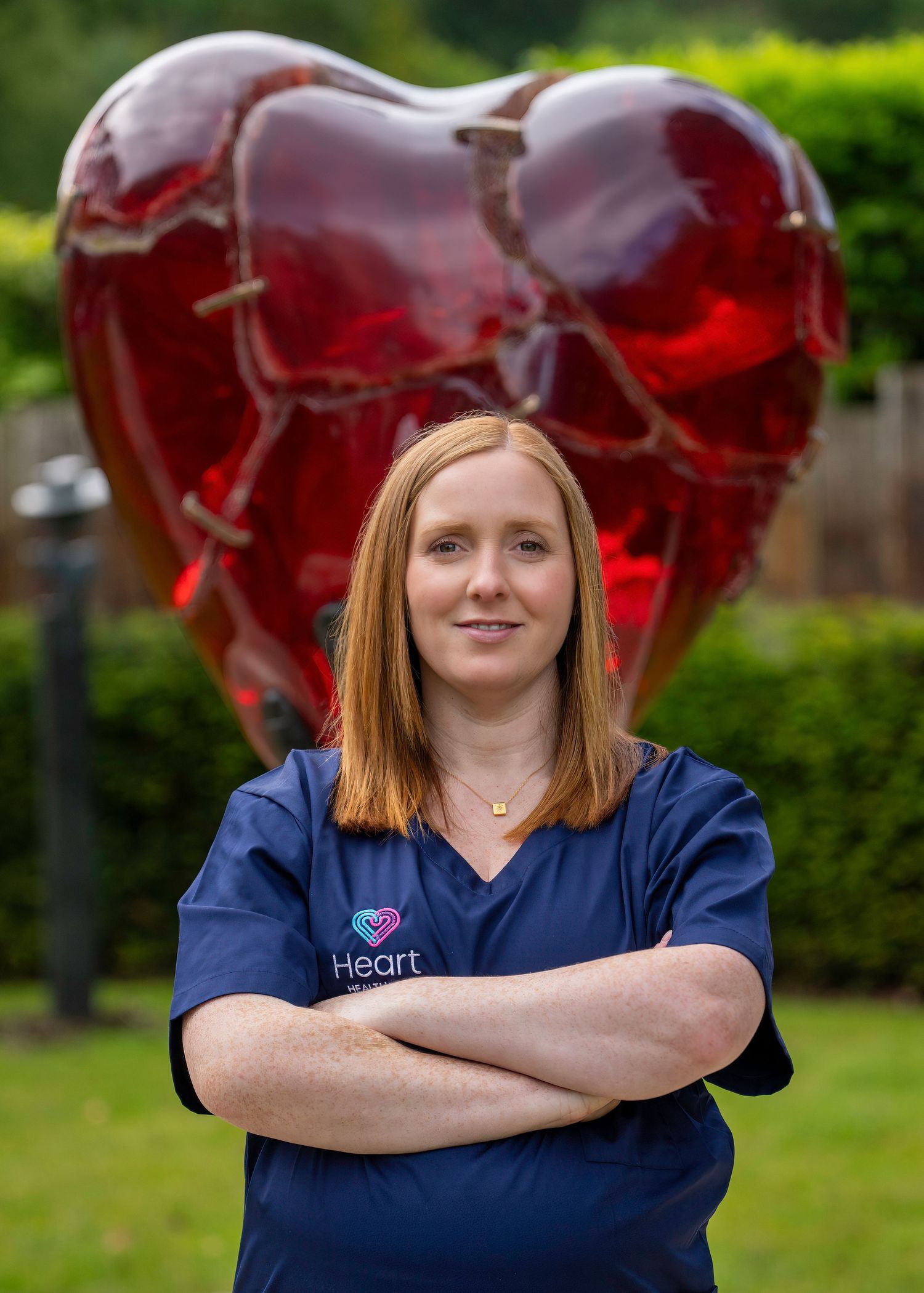 Woman in navy Heart medical scrubs standing with arms crossed in front of a large red heart sculpture. Dave Martin Photography Belfast