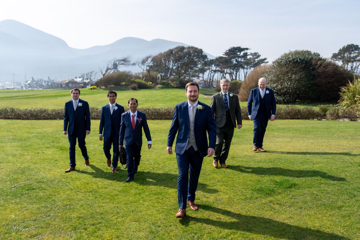 Six groomsmen in navy suits walking confidently across a green lawn with scenic mountains in the background. Slieve Donard Hotel