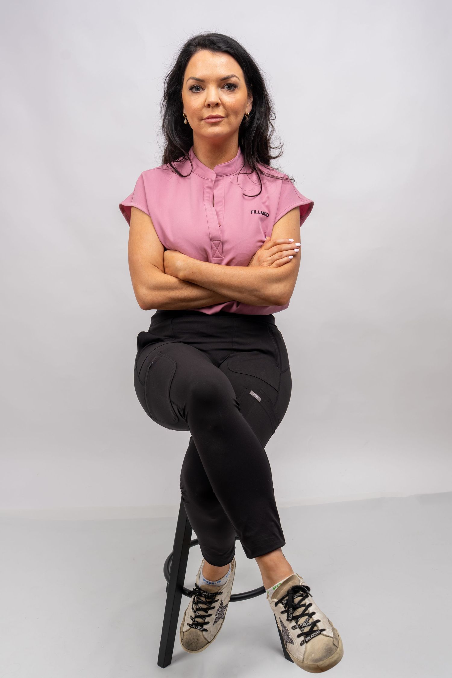 Woman in pink polo shirt and black pants sits cross-armed on stool, wearing sneakers, against gray background. Dave Martin Photography