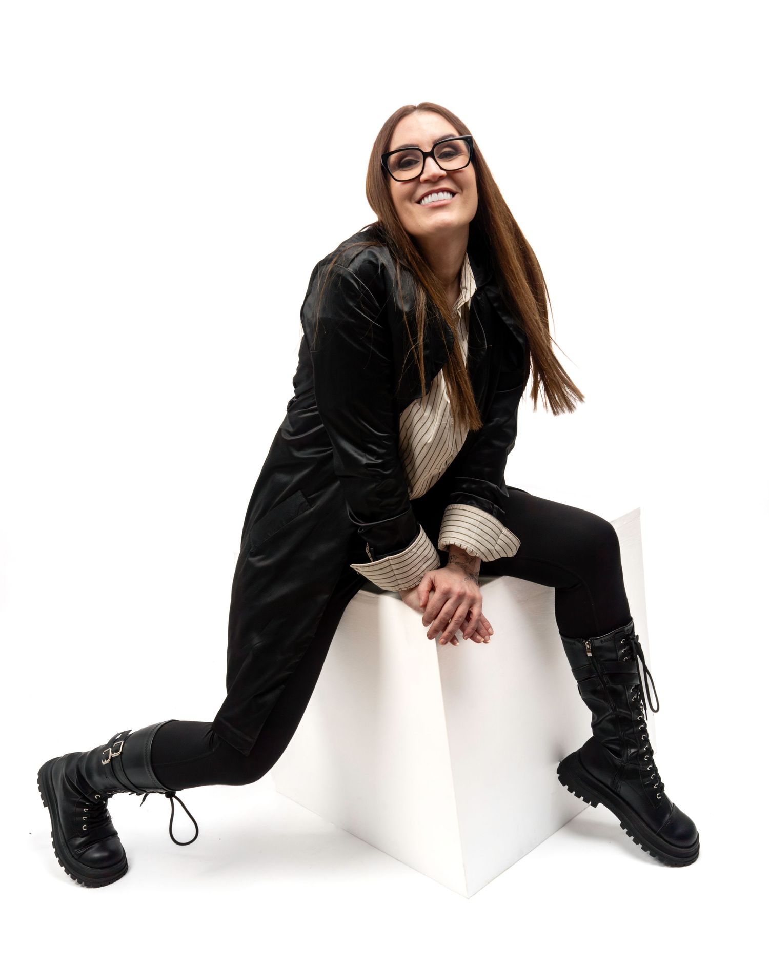 Smiling brunette woman in black coat and boots sitting on a white cube against white background. Dave Martin Photography Studio Belfast