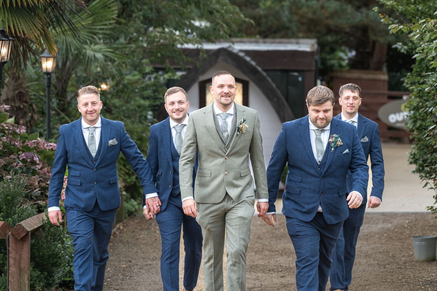 Groom in sage suit leads groomsmen in navy suits walking confidently at outdoor wedding venue. Dave Martin Photographer Northern Ireland