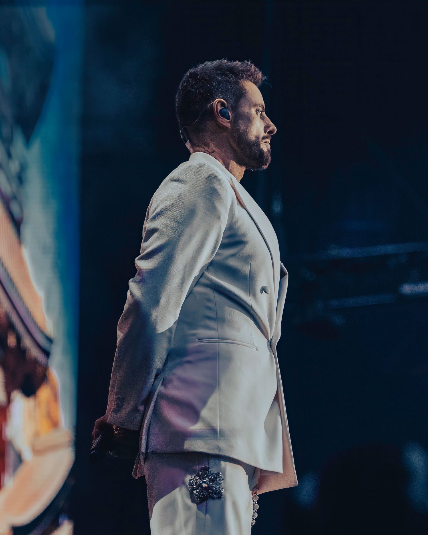 Male performer in white double-breasted suit stands in profile on dimly lit concert stage. Dave Martin celebrity Photography