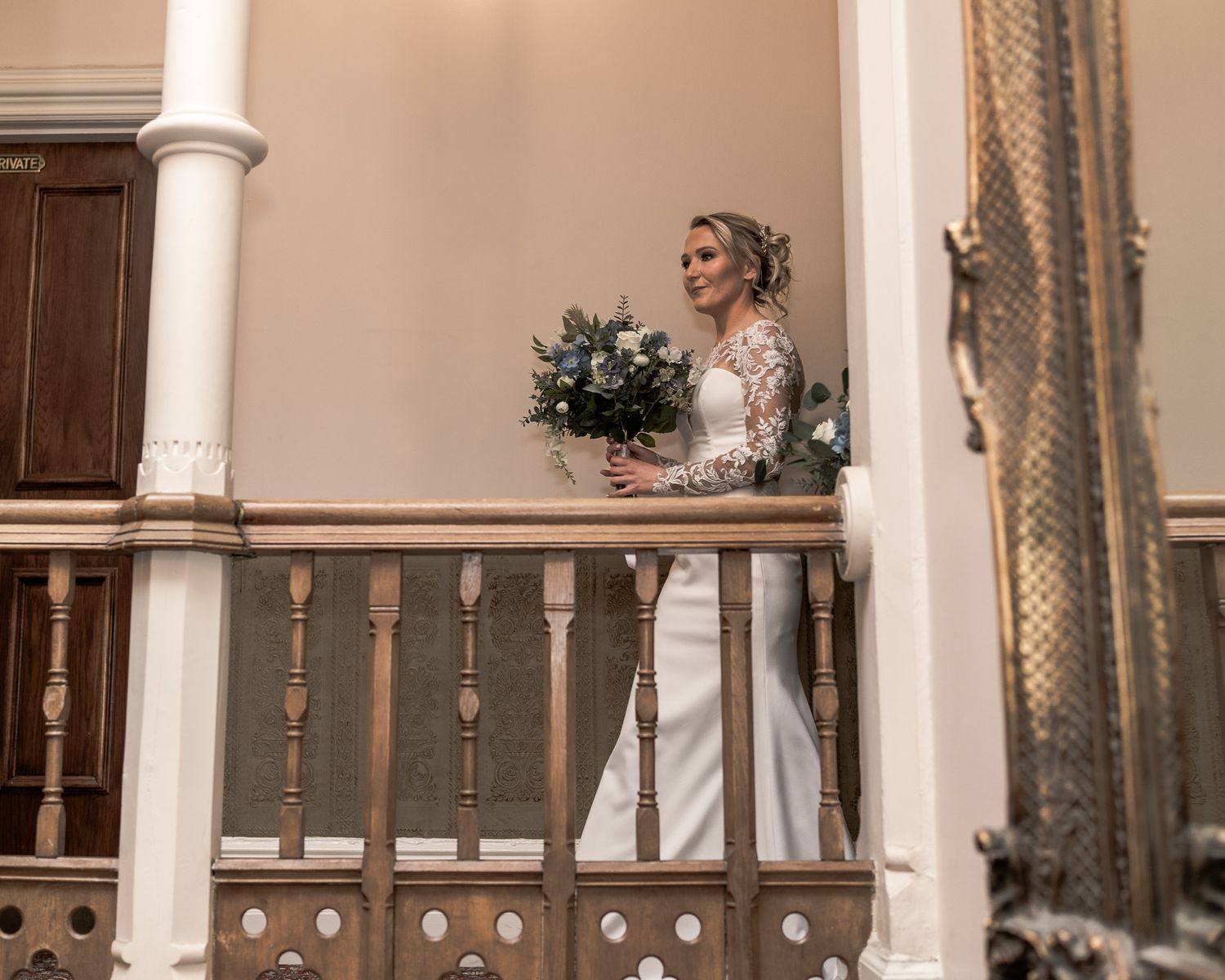 Bride in white lace gown holding a bouquet stands on an elegant wooden staircase in a grand venue.