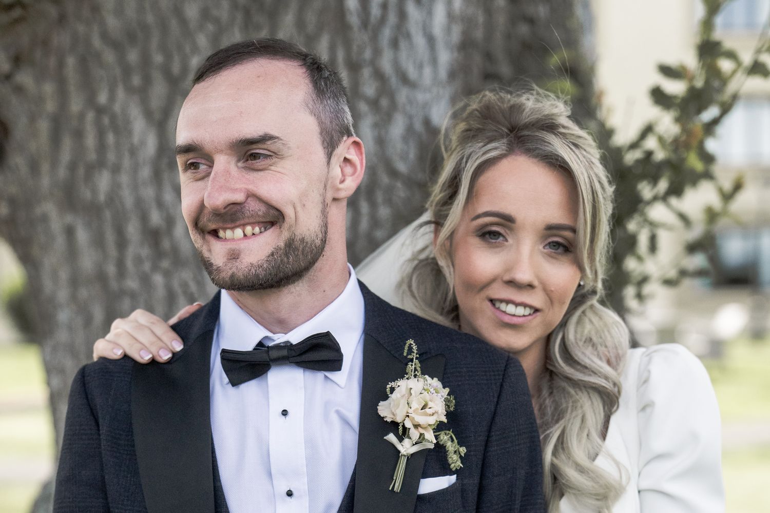 Happy couple posing outdoors on their wedding day, groom in tuxedo and bride in white dress with floral boutonniere. Northern ireland